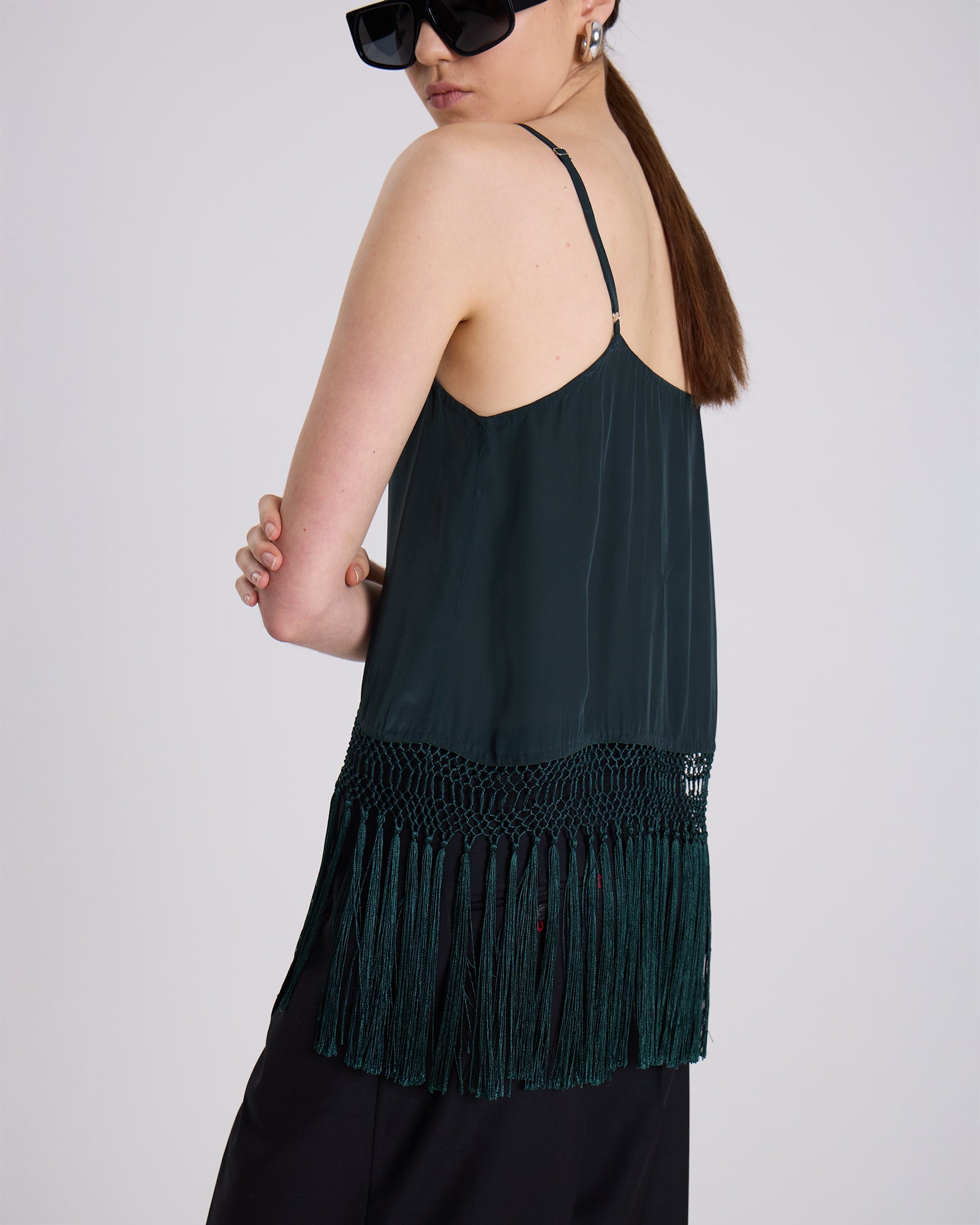 Person wearing a dark green tank top with fringe details on a plain background