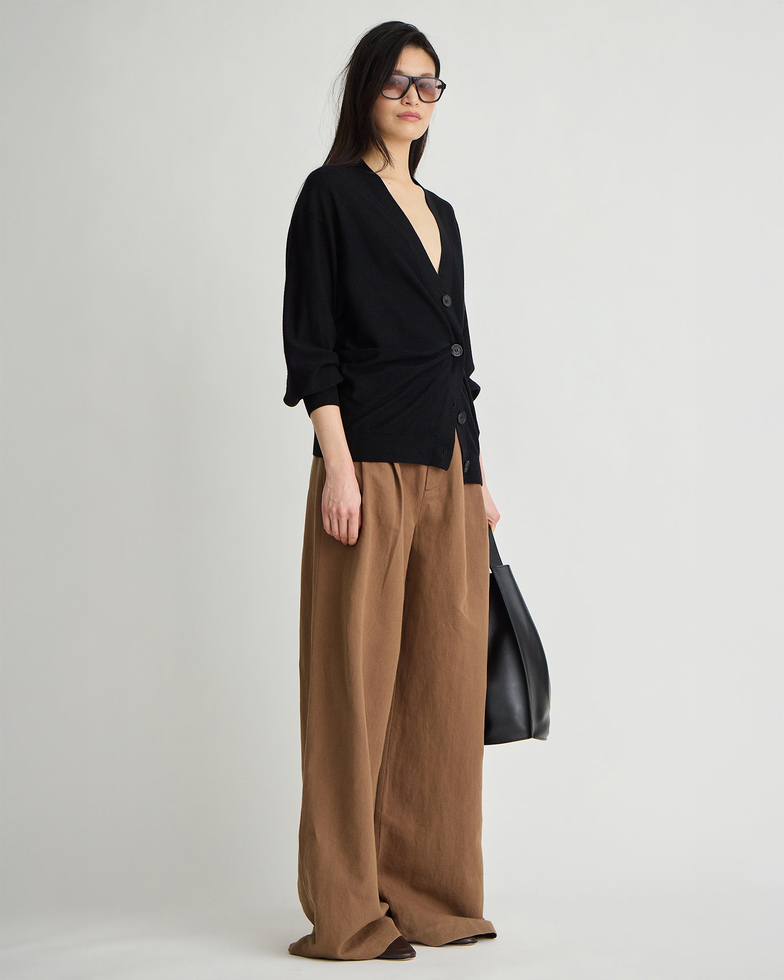 model wearing tobacco pleated pants and a black cardigan and sunglasses from the side