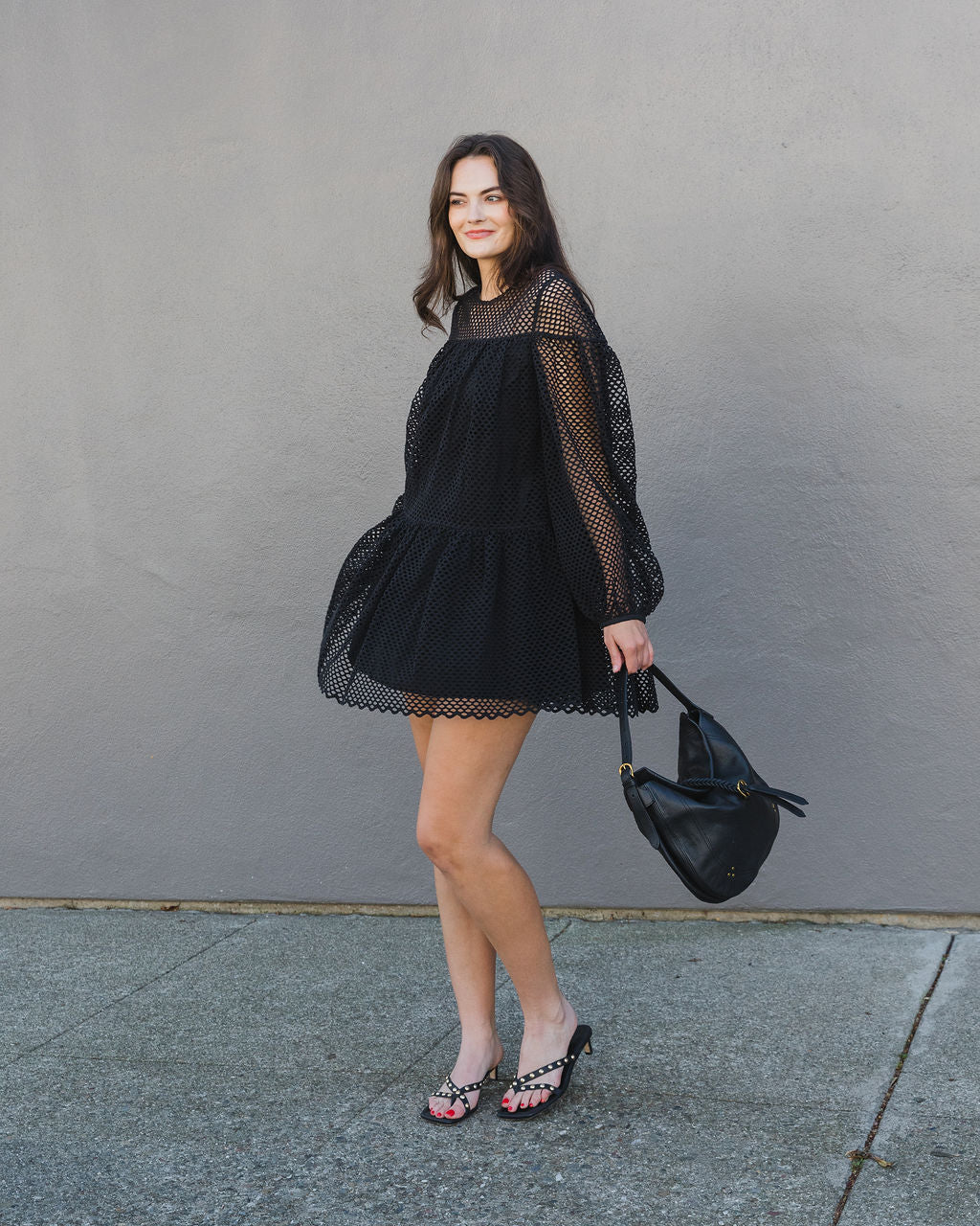 Woman wearing a black dress with sheer sleeves against a gray wall.