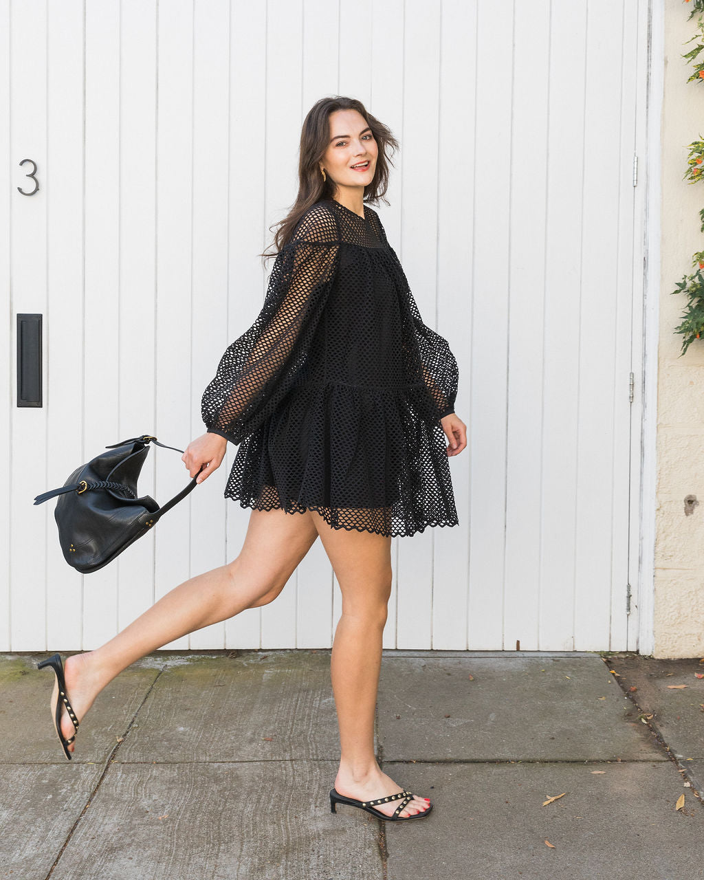 Woman in a black lace dress holding a black handbag against a white door background