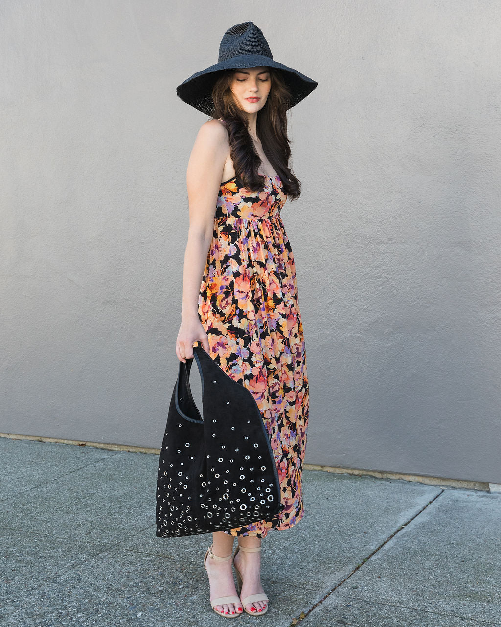 Woman in a floral dress and wide-brimmed hat holding a black bag against a gray wall.