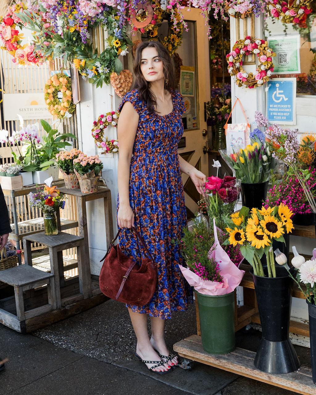 Woman in a floral dress standing in front of a flower shop with various flowers and plants.
