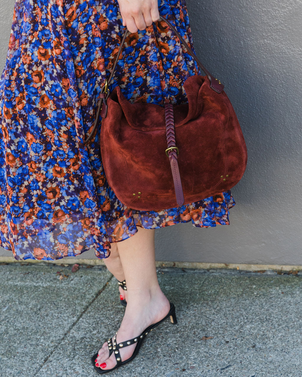 Person wearing a floral dress and holding a brown suede handbag against a gray wall.