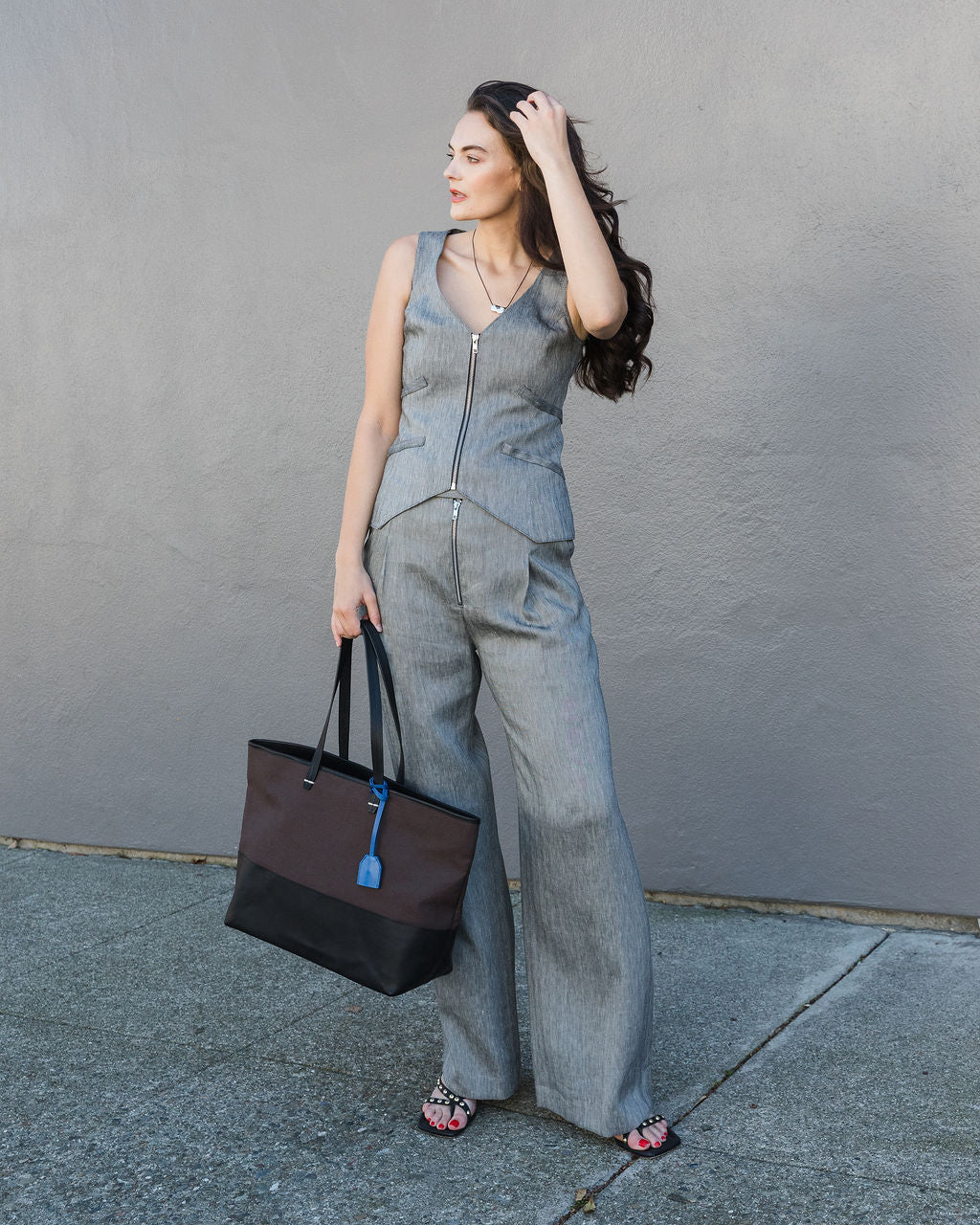 Woman in a denim outfit holding a black tote bag against a gray wall.