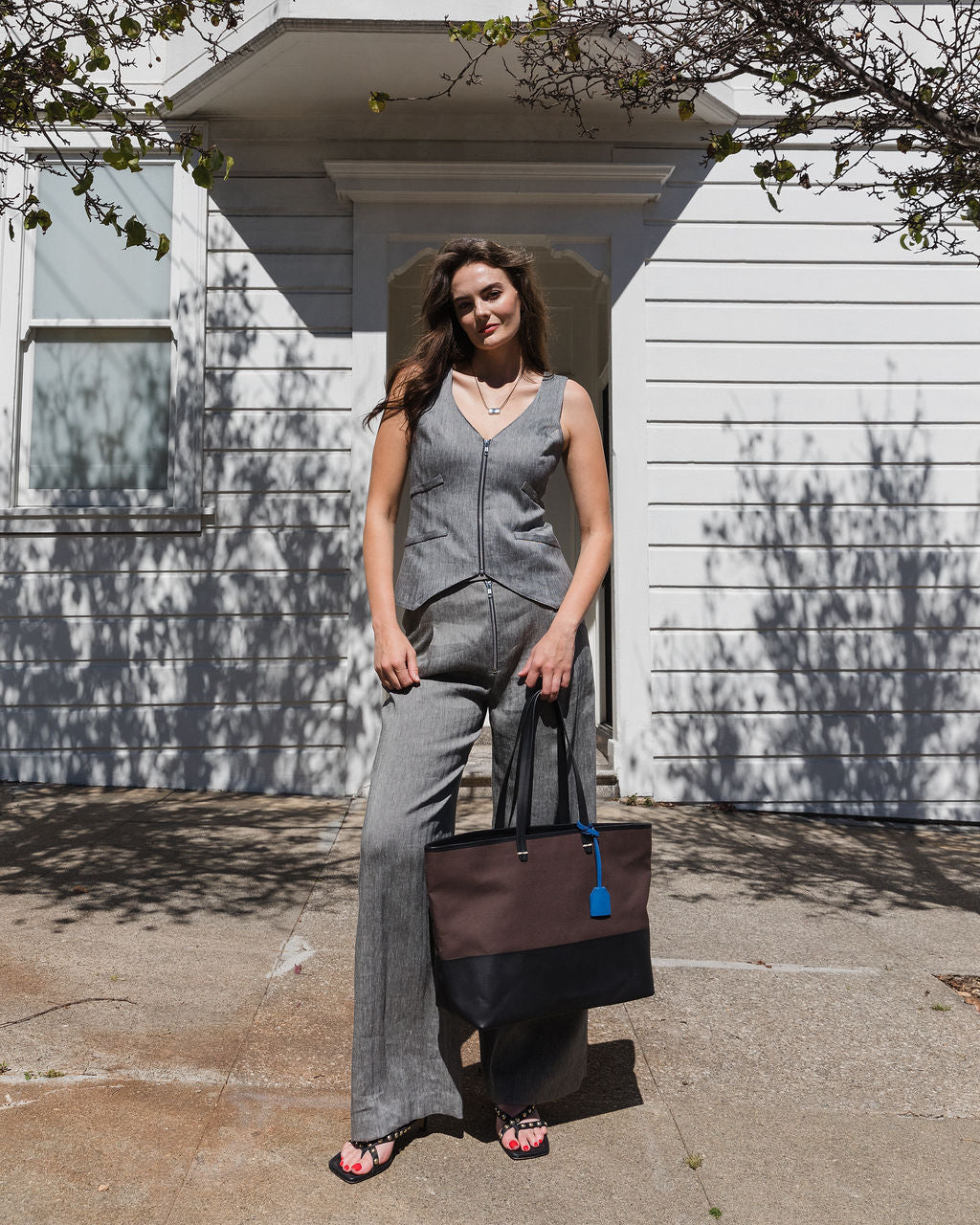 Woman in a gray outfit standing outside a house holding a black bag.
