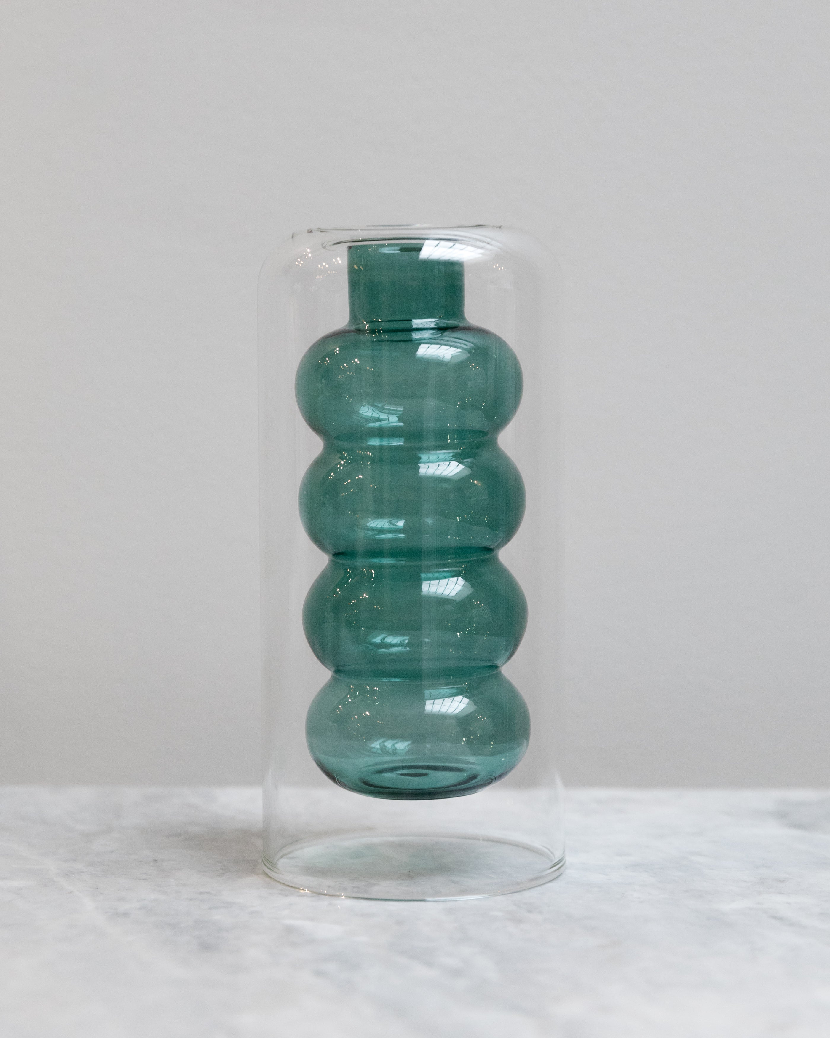 Green glass vase with ribbed design inside a clear glass container on a light gray background
