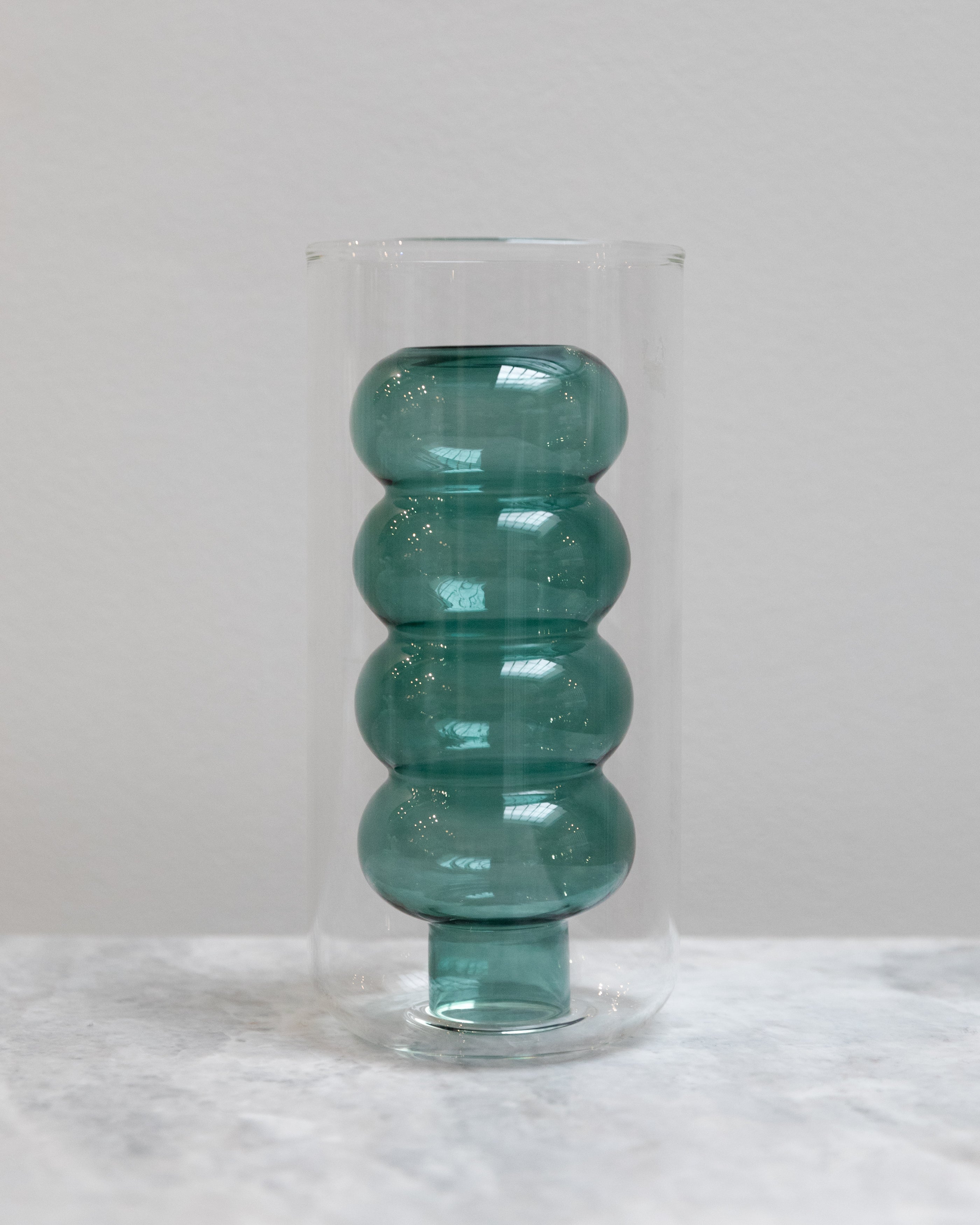 Teal glass vase with ribbed design inside a clear glass cover on a light gray background