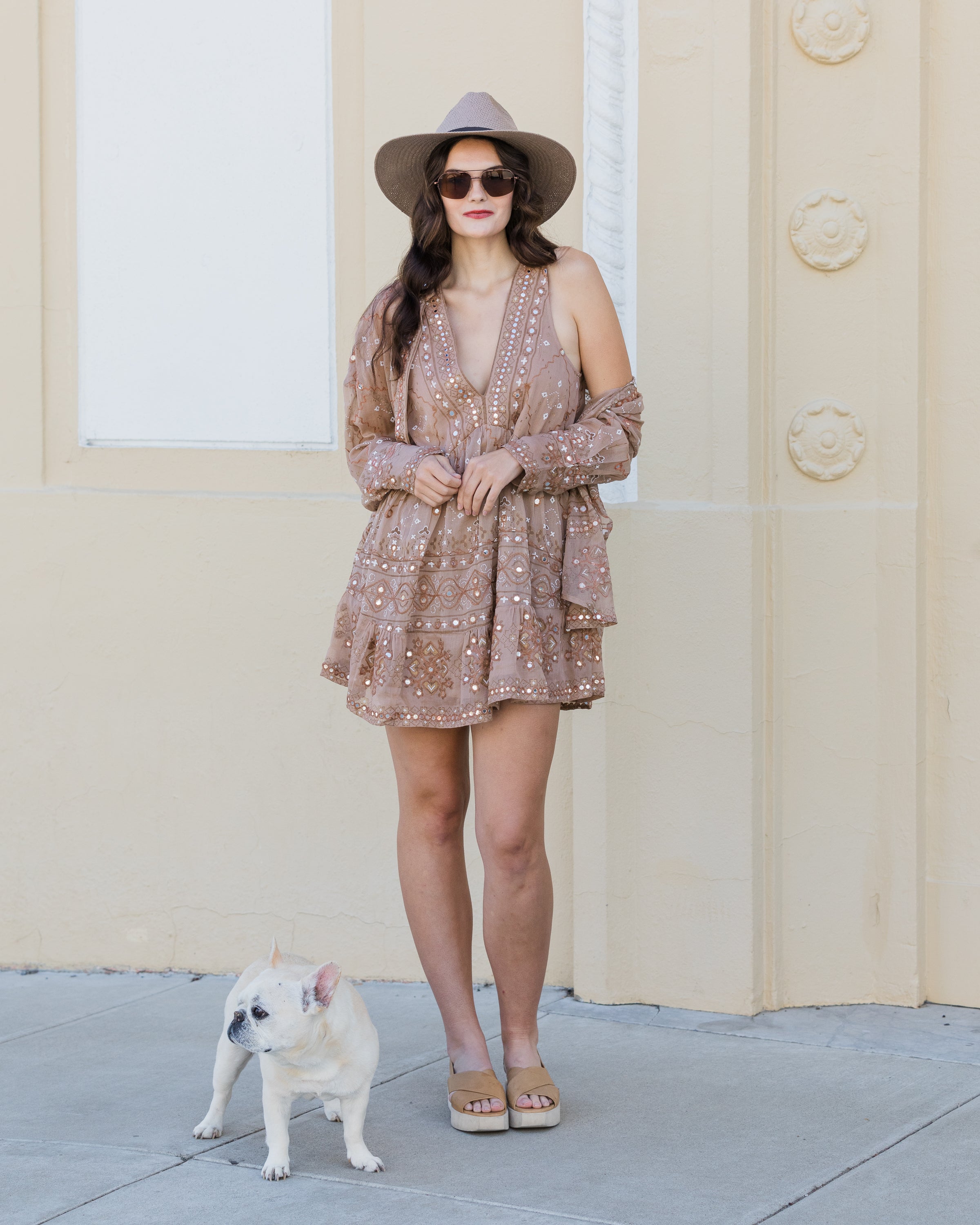 Woman in a patterned dress and hat standing next to a small dog on a sidewalk.