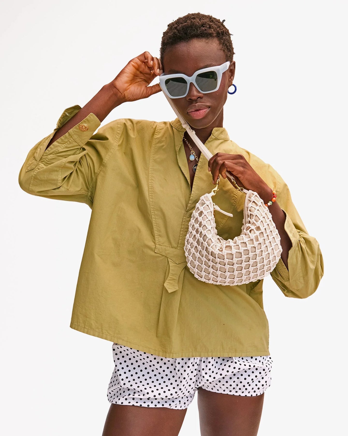 Woman in a green jacket and polka dot shorts holding a white handbag against a white background