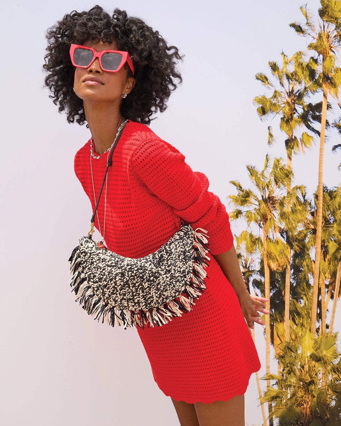 Woman wearing a red outfit with a textured bag, standing against a light background with plants.