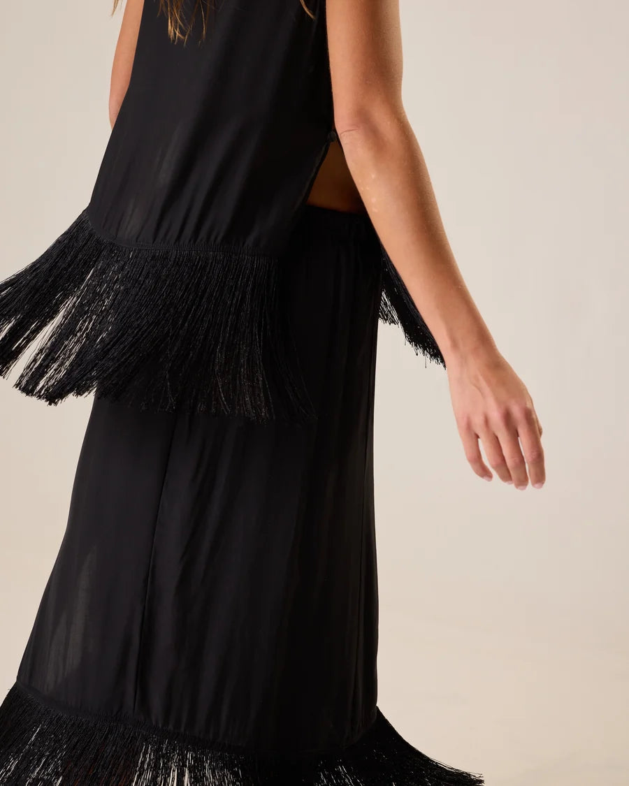A woman wearing a sleeveless black dress with layered fringe trim, shown from the back against a plain light beige studio background.