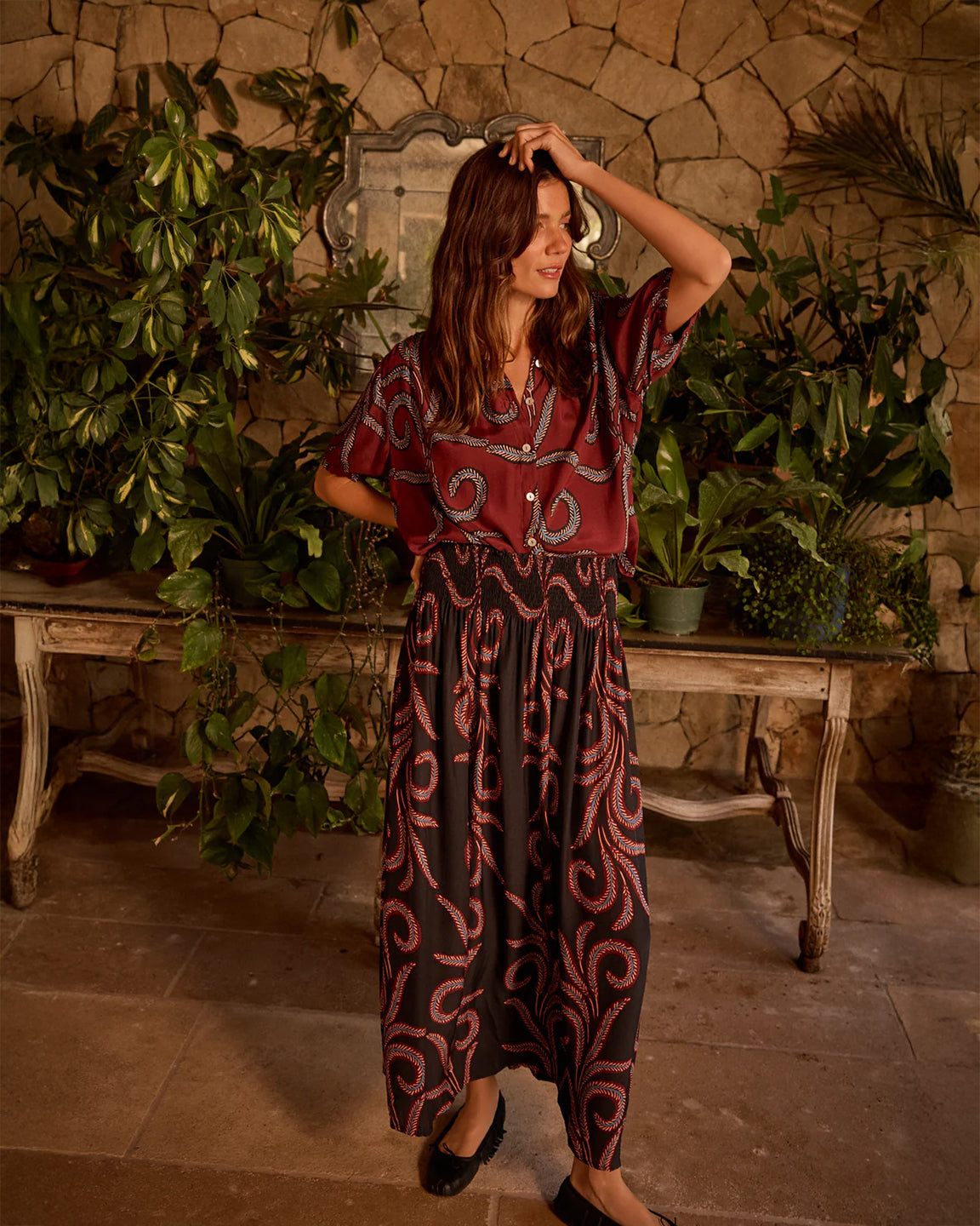 Woman in a patterned dress standing in a rustic indoor setting with plants and stone walls.