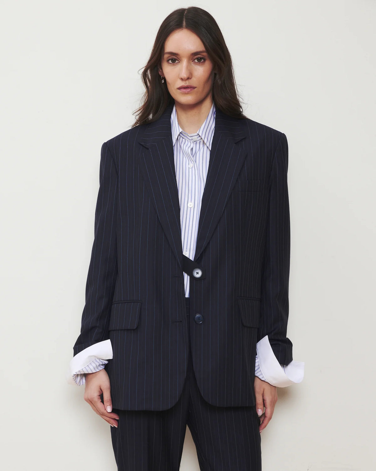 A woman modeling an oversized dark navy pinstripe blazer with matching trousers, styled over a white and blue striped button-down shirt, against a plain light beige background.