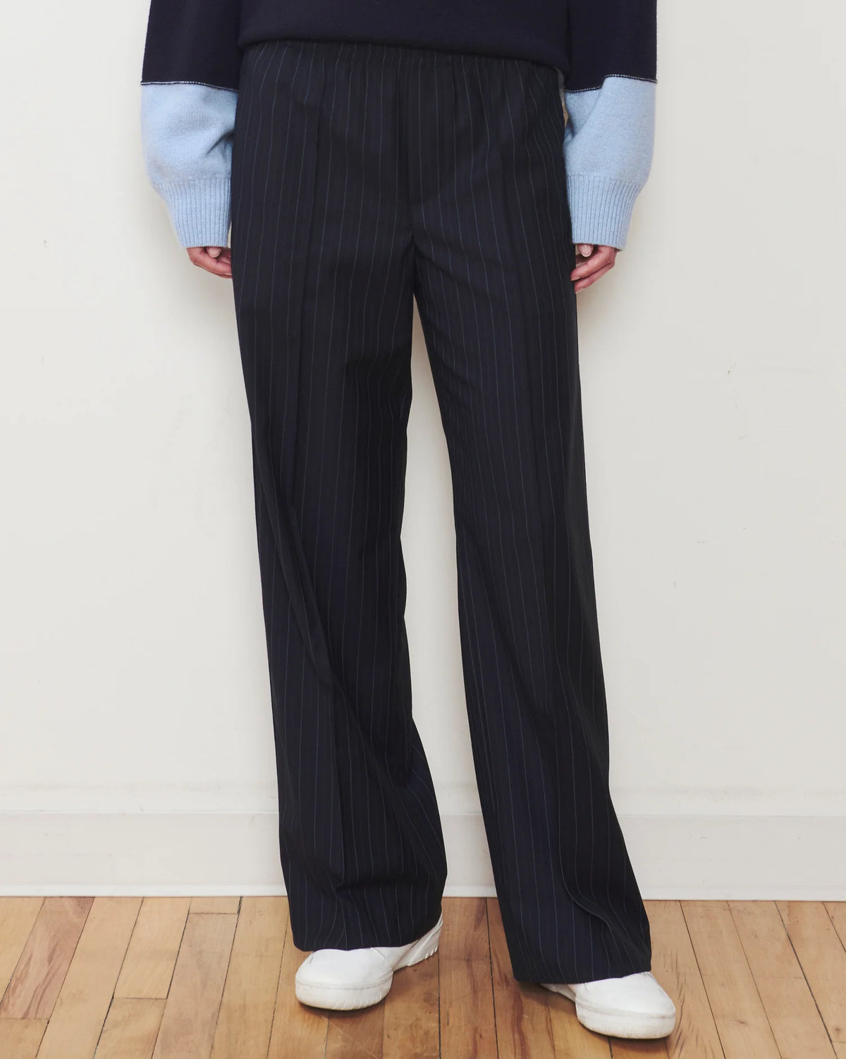 A person wearing dark navy pinstripe wide-leg trousers, a dark and light blue colorblock sweater, and white sneakers, standing against a white wall on a wooden floor.
