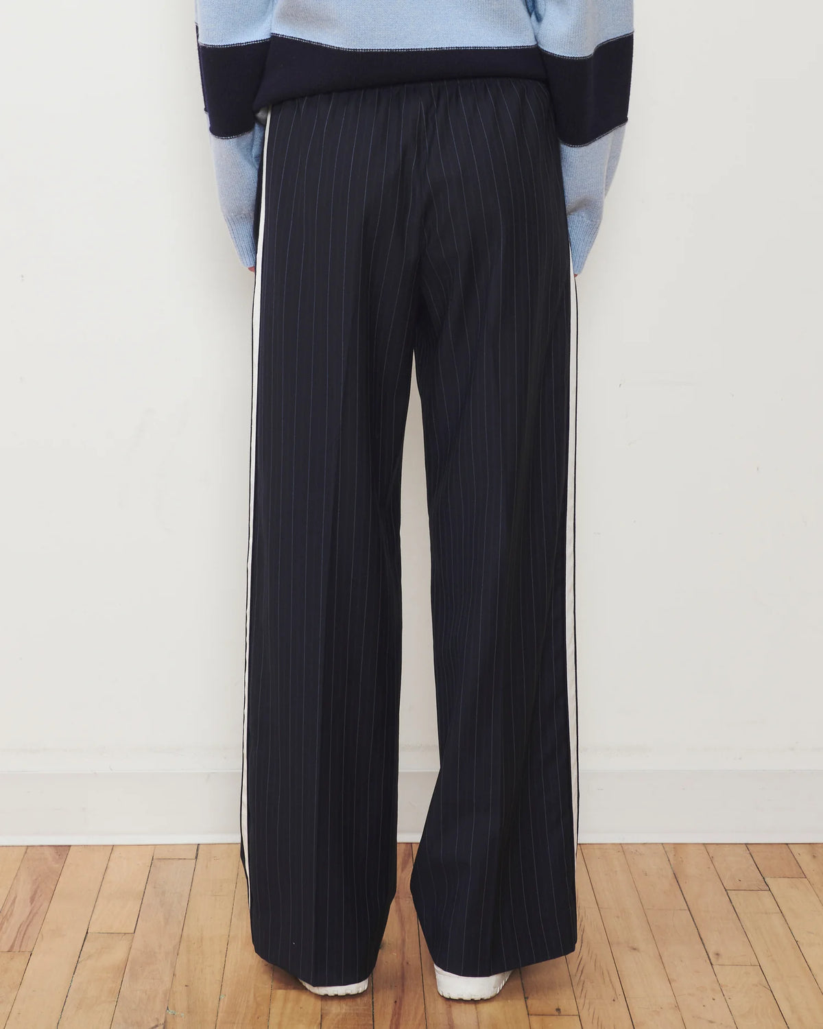 A person shown from the back wearing wide-leg pleated trousers in a very dark navy or black with thin vertical white pinstripes, paired with a light blue and navy color-blocked sweater. The setting is indoors with a white wall and light wood floor.