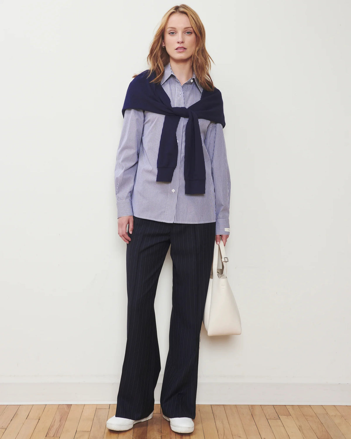 A woman stands against a plain off-white wall wearing a light blue and white striped button-up shirt, dark navy wide-leg trousers, a navy knit wrap tied around her shoulders, and white sneakers. She holds a cream-colored handbag.