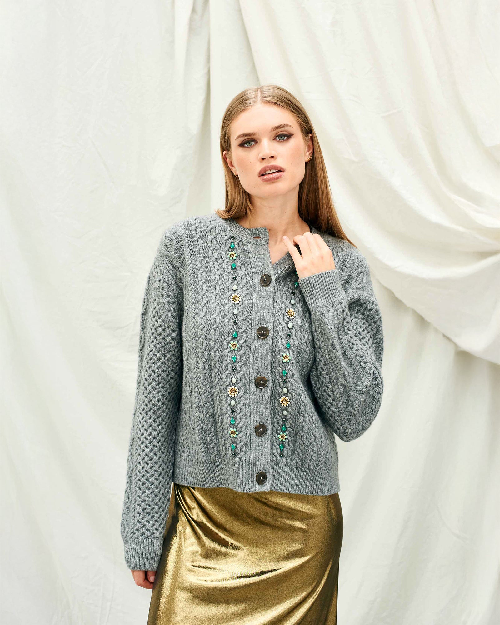 Woman wearing a gray knitted cardigan with decorative buttons against a white curtain background