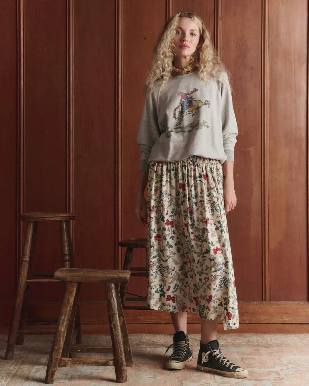 Woman wearing a patterned skirt and sweater standing in a room with wooden paneling.