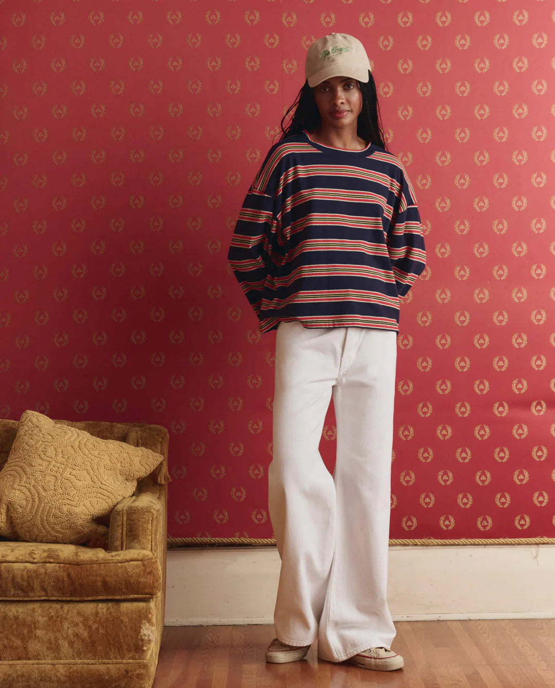 Person wearing a striped sweater and white pants standing against a patterned red wall.