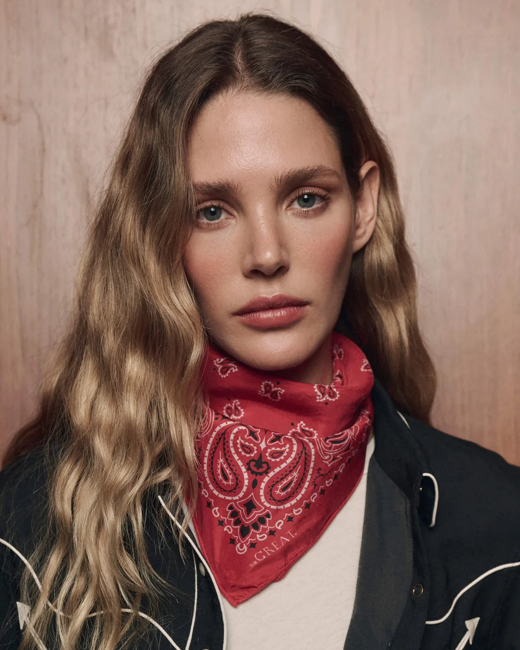 A close-up portrait of a woman with long wavy blonde hair, wearing a red paisley bandana around her neck and a dark jacket over a cream shirt, set against a warm wood-textured background.