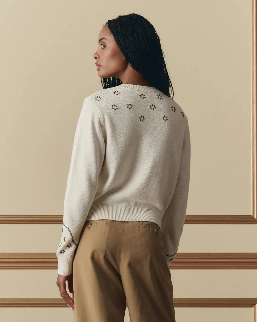 Woman wearing a beige sweater with floral embellishments and beige pants against a beige wall.