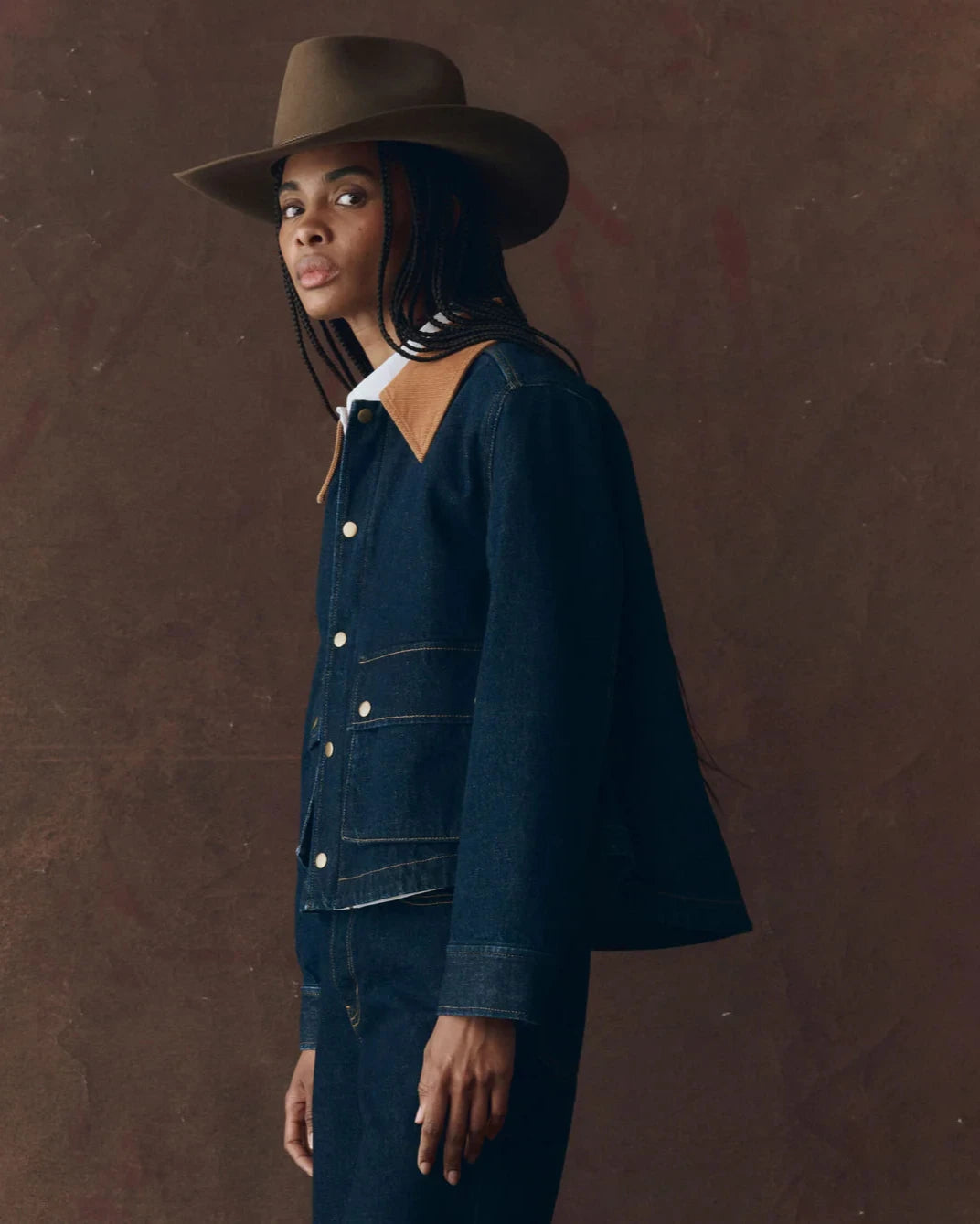 Woman wearing a dark denim jacket and hat against a brown background