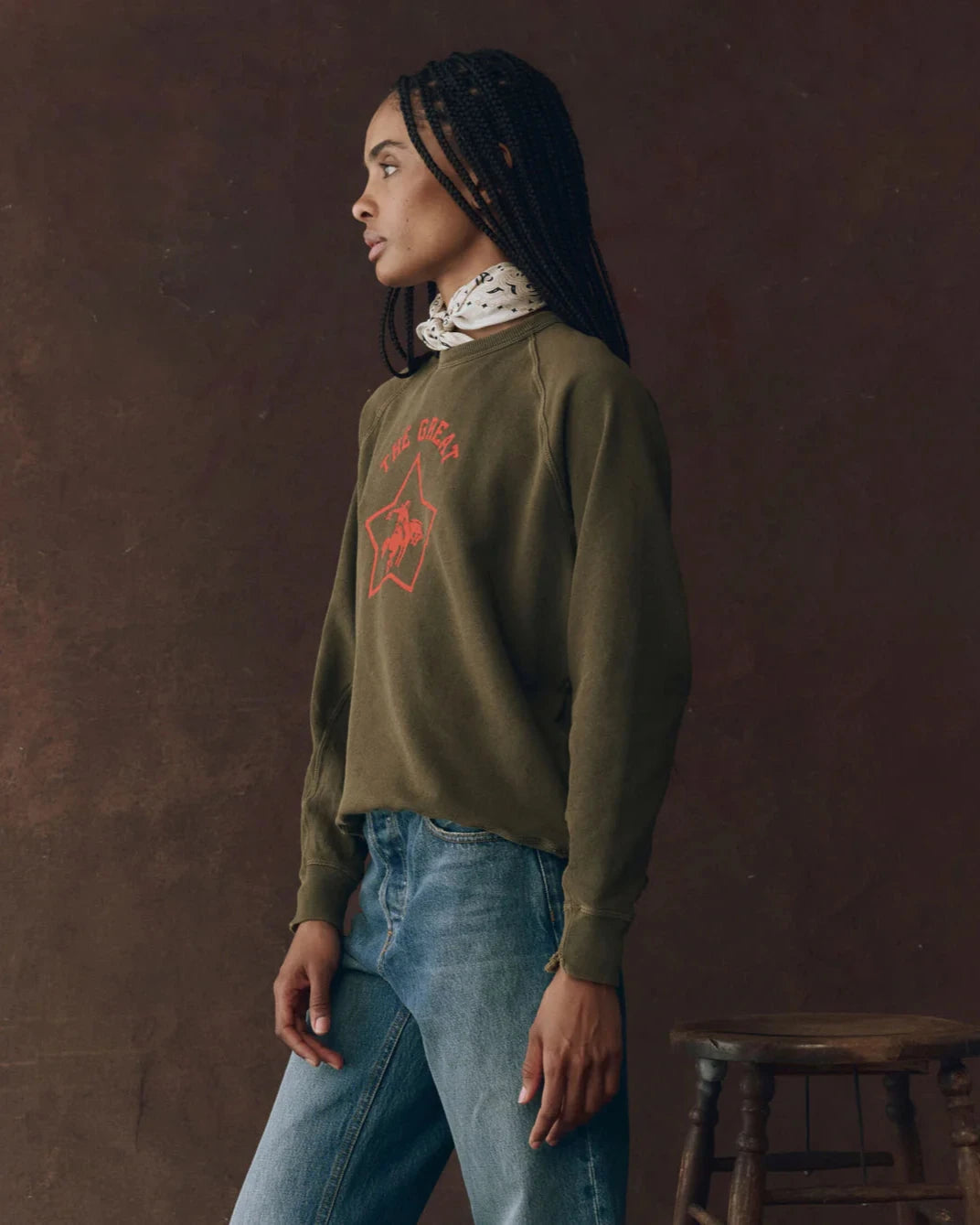 Person wearing a green sweatshirt with a red logo and blue jeans against a brown background