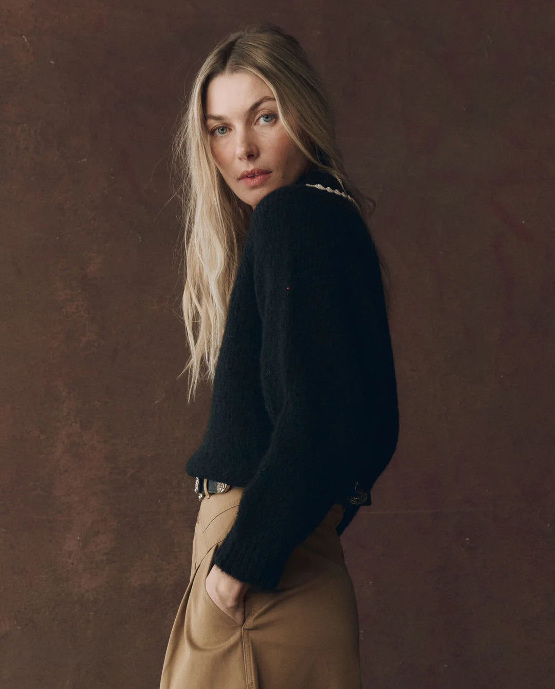 Woman wearing a black sweater and beige pants against a brown background