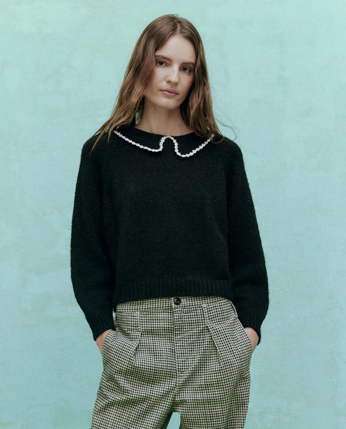 A woman wearing a black pullover sweater with a crochet collar and soft white contrast scallop trim, paired with patterned trousers.