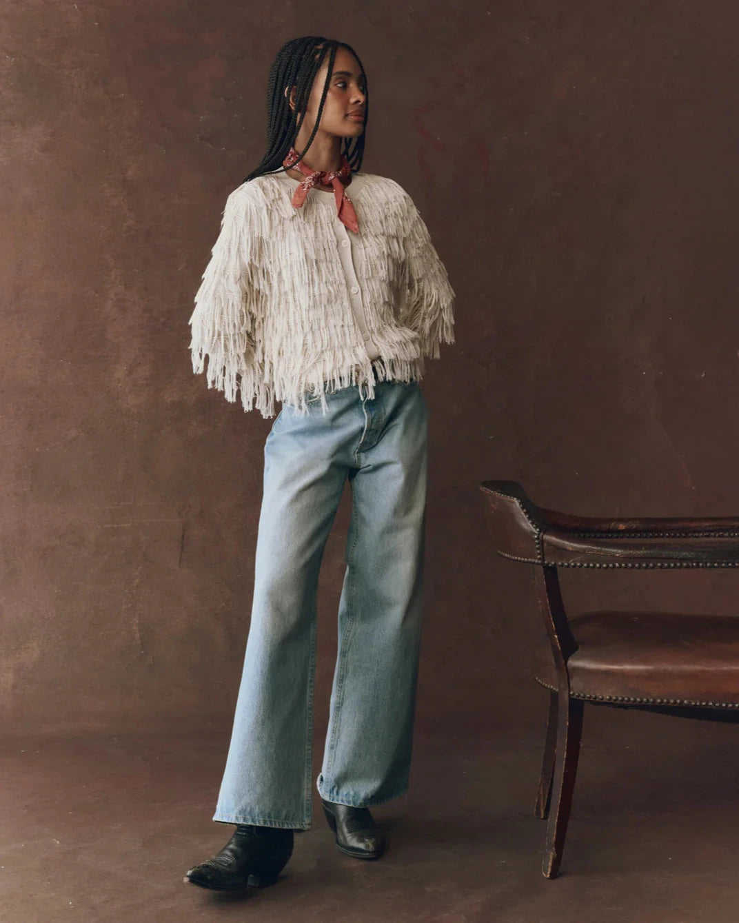 Woman wearing a white fringed sweater and light blue jeans standing against a brown background.