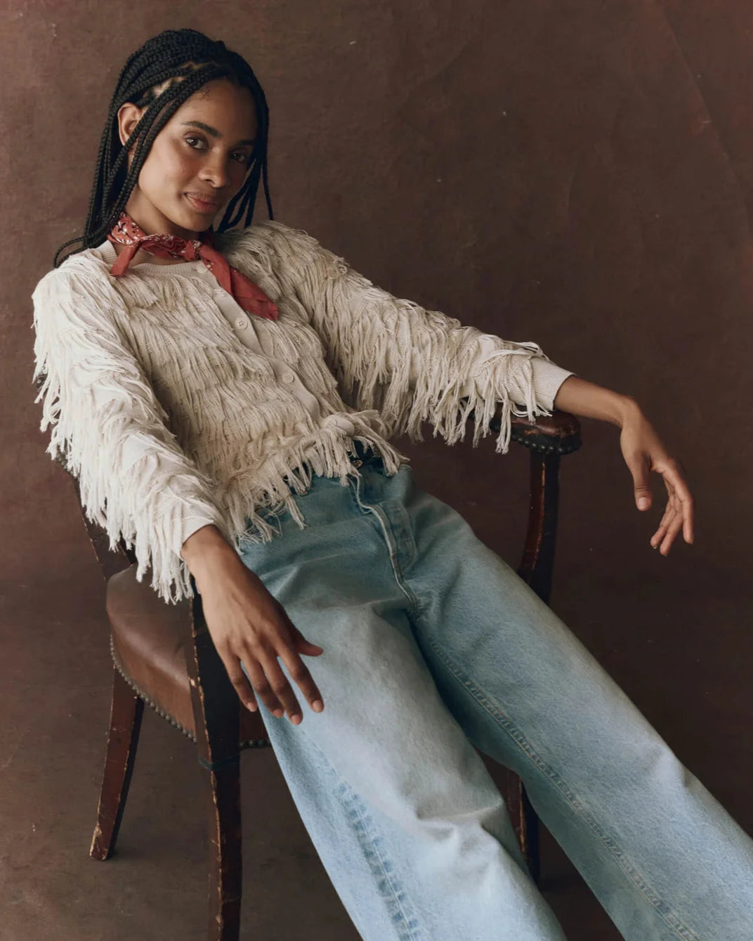 Woman sitting on a chair wearing a fringed sweater and light blue jeans against a brown background