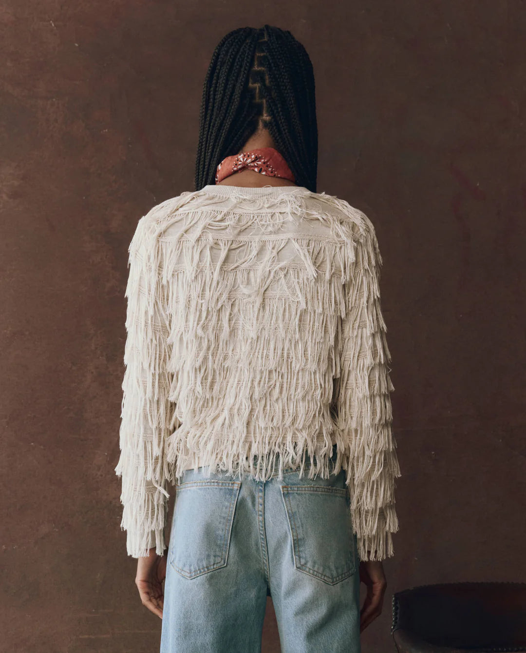 Person wearing a beige fringed jacket and light blue jeans against a brown background