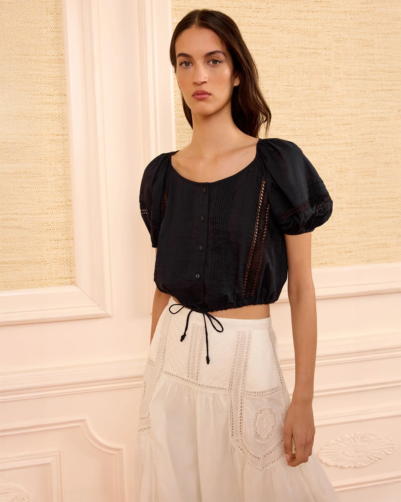 A woman modeling a black puff-sleeve crop top with side cutouts and a white embroidered skirt, standing against a cream-colored paneled wall.