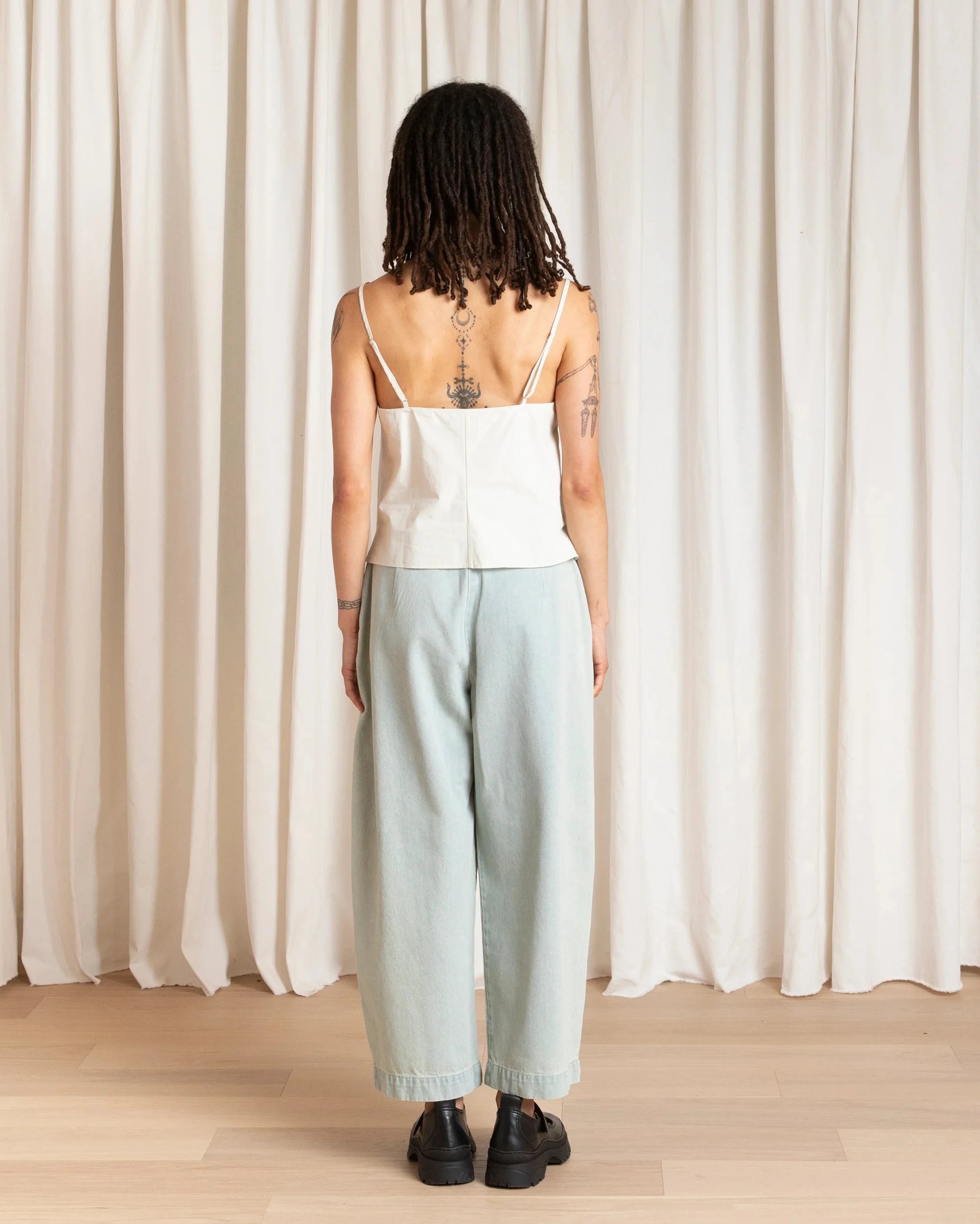 Person wearing a white top and light blue pants standing in front of a white curtain.