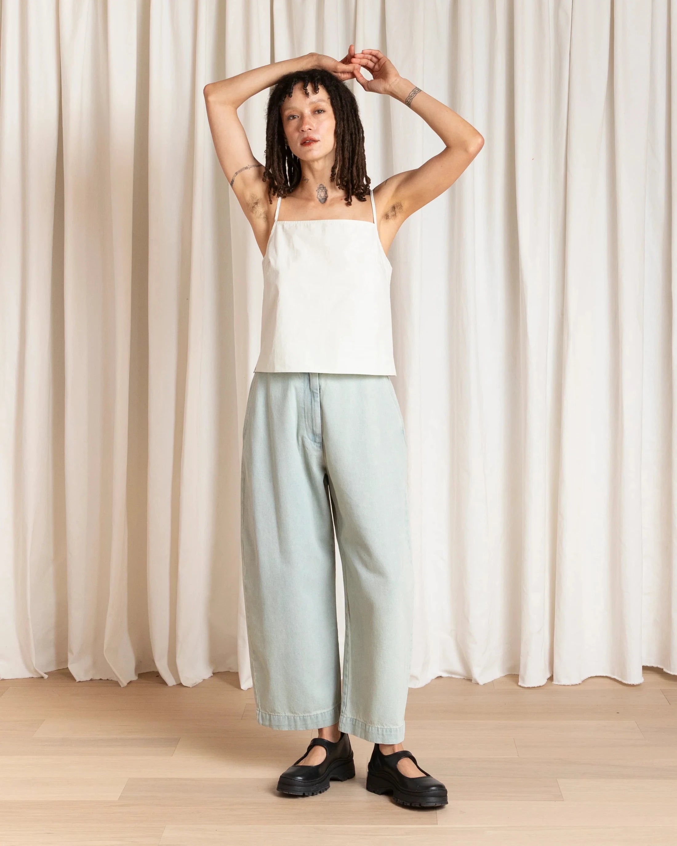 Woman wearing a white top and light blue pants standing against a white curtain.