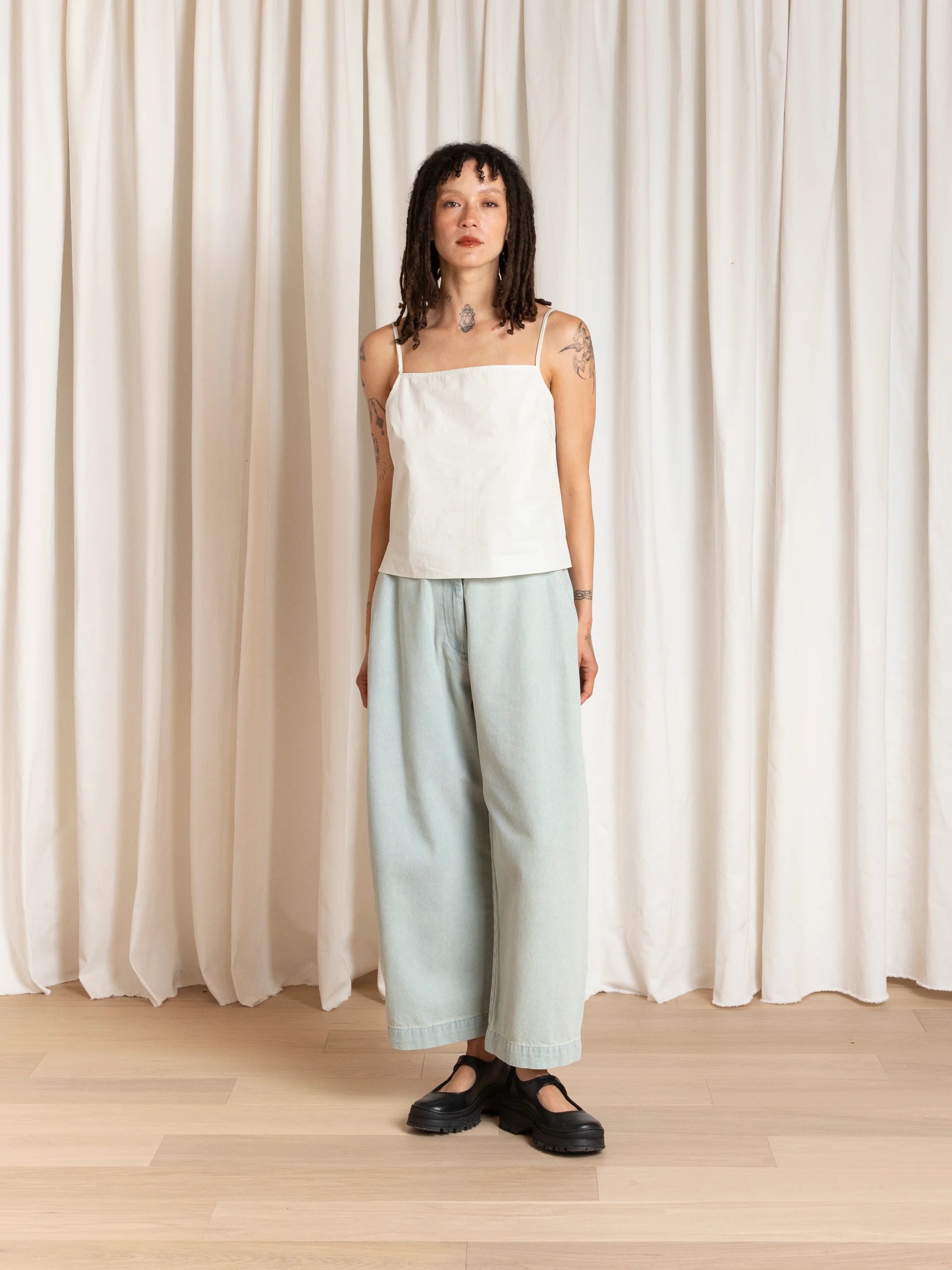 Woman wearing a white top and light blue pants standing in front of white curtains.