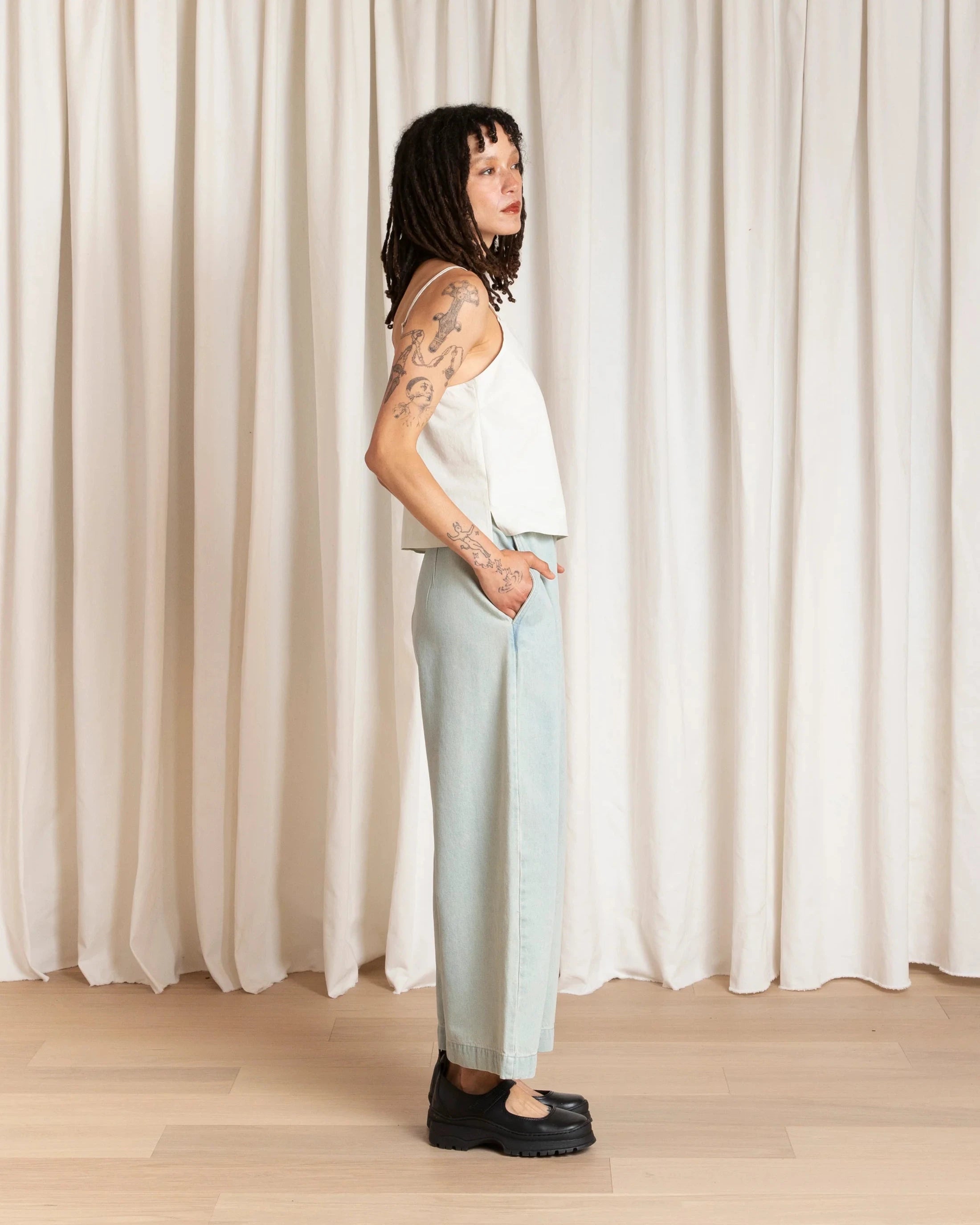 Person wearing a white sleeveless top and light blue pants standing against a white curtain.