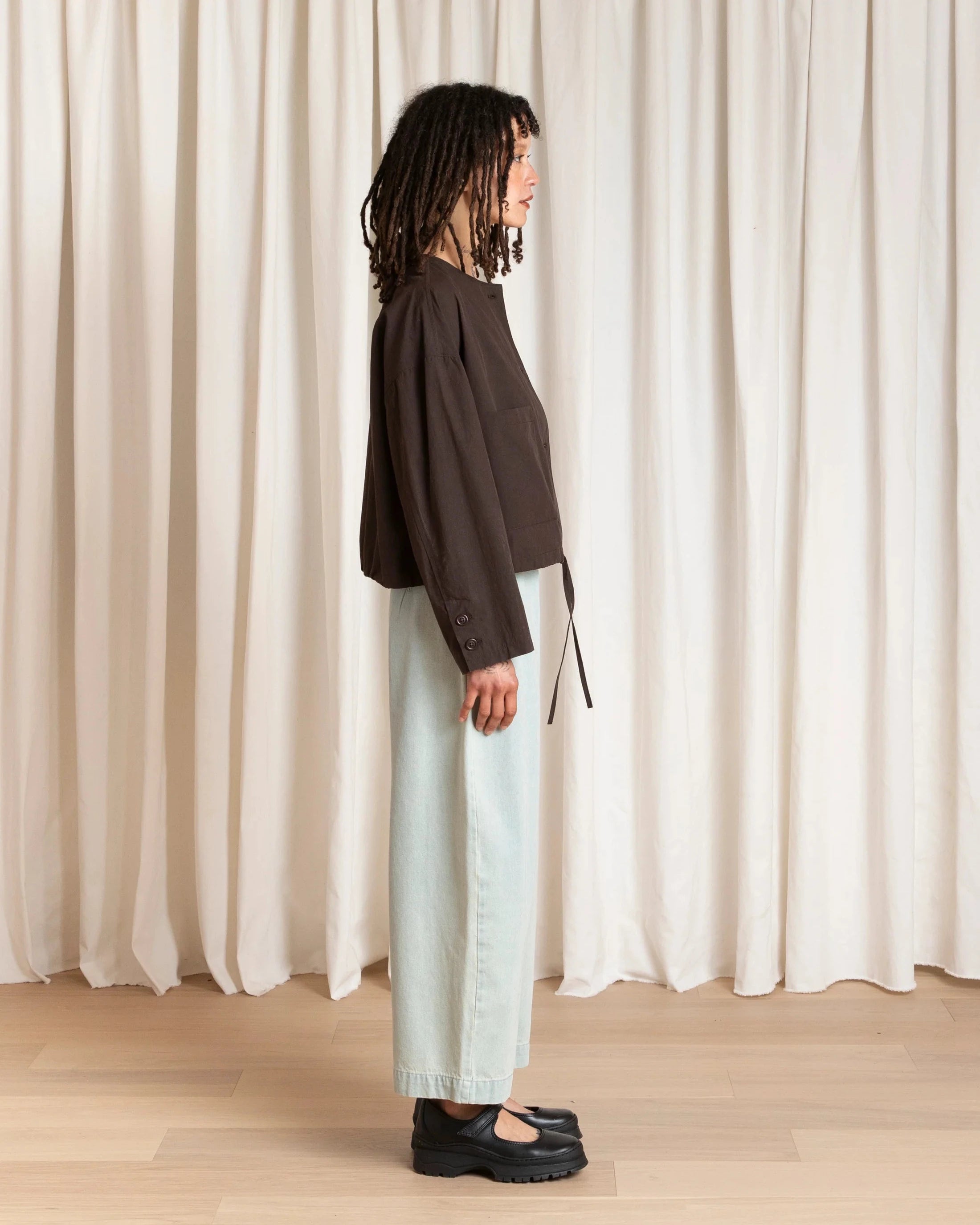 Person wearing a dark brown top and light blue jeans standing against a white curtain.