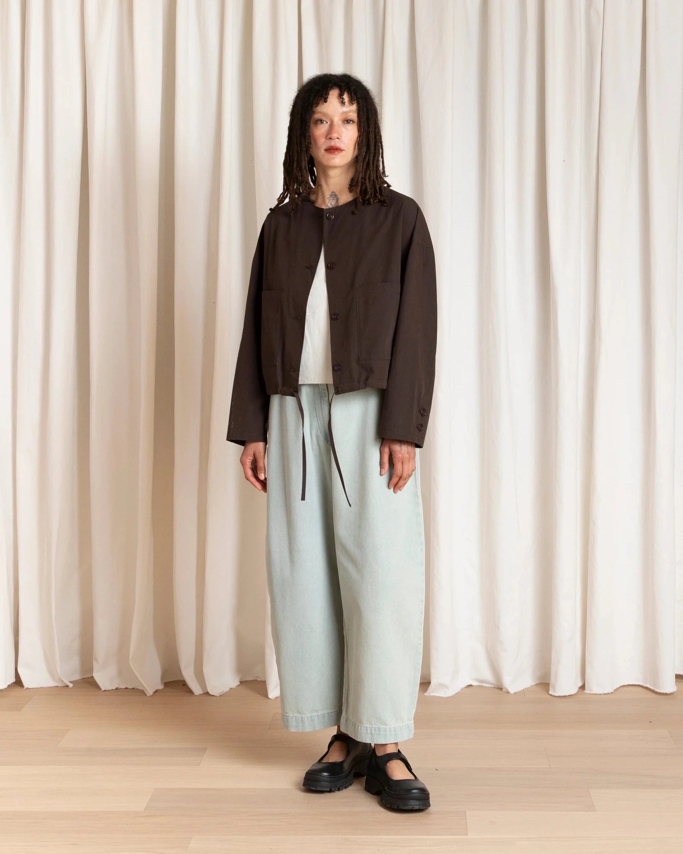 Person wearing a dark jacket and light pants standing against a white curtain.