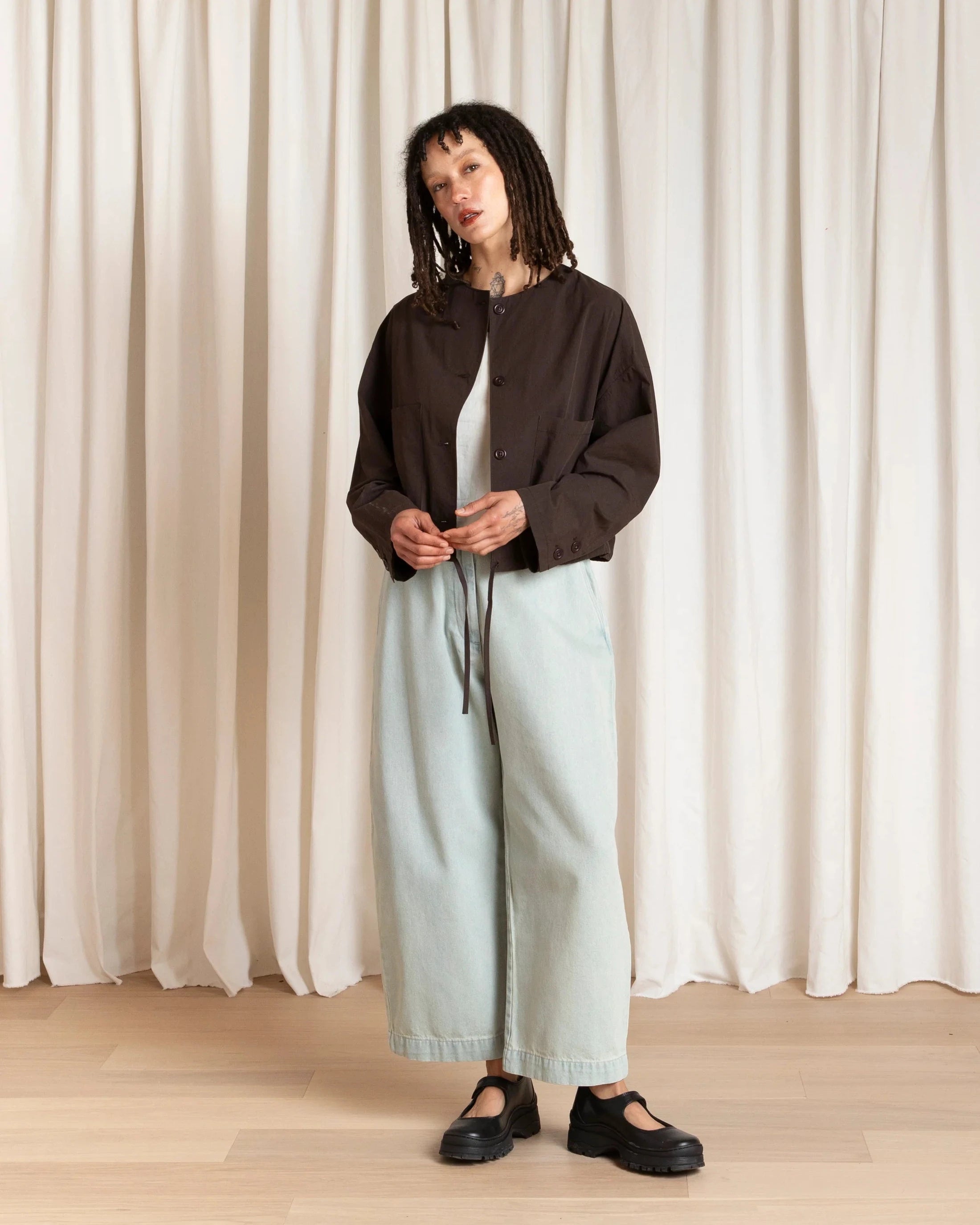 Person wearing a dark jacket, light blue pants, and black shoes standing against a white curtain.