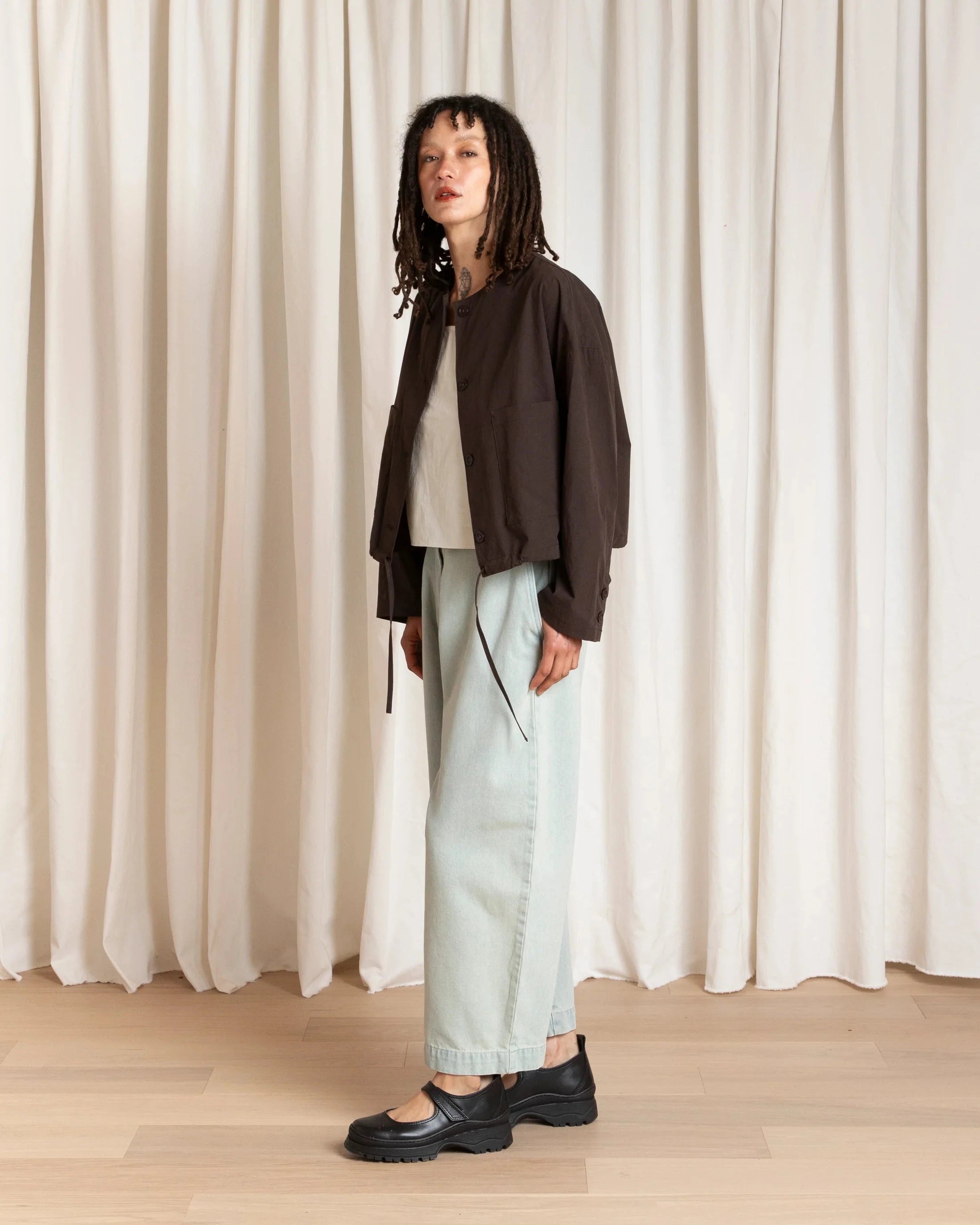 Person wearing a brown jacket, white top, light green pants, and black shoes standing against a white curtain.