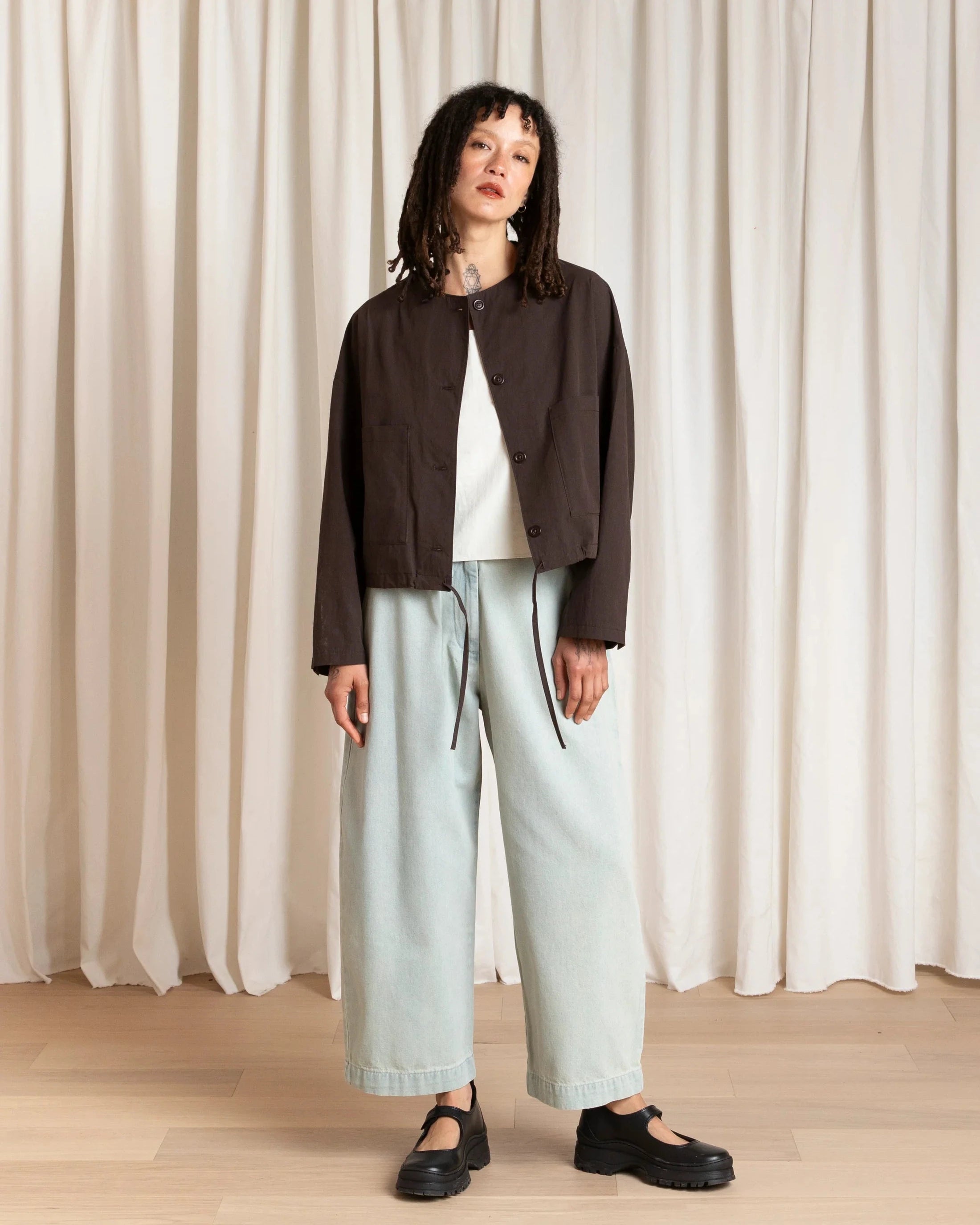 Woman wearing a brown jacket, white top, light blue pants, and black shoes against a white curtain background.