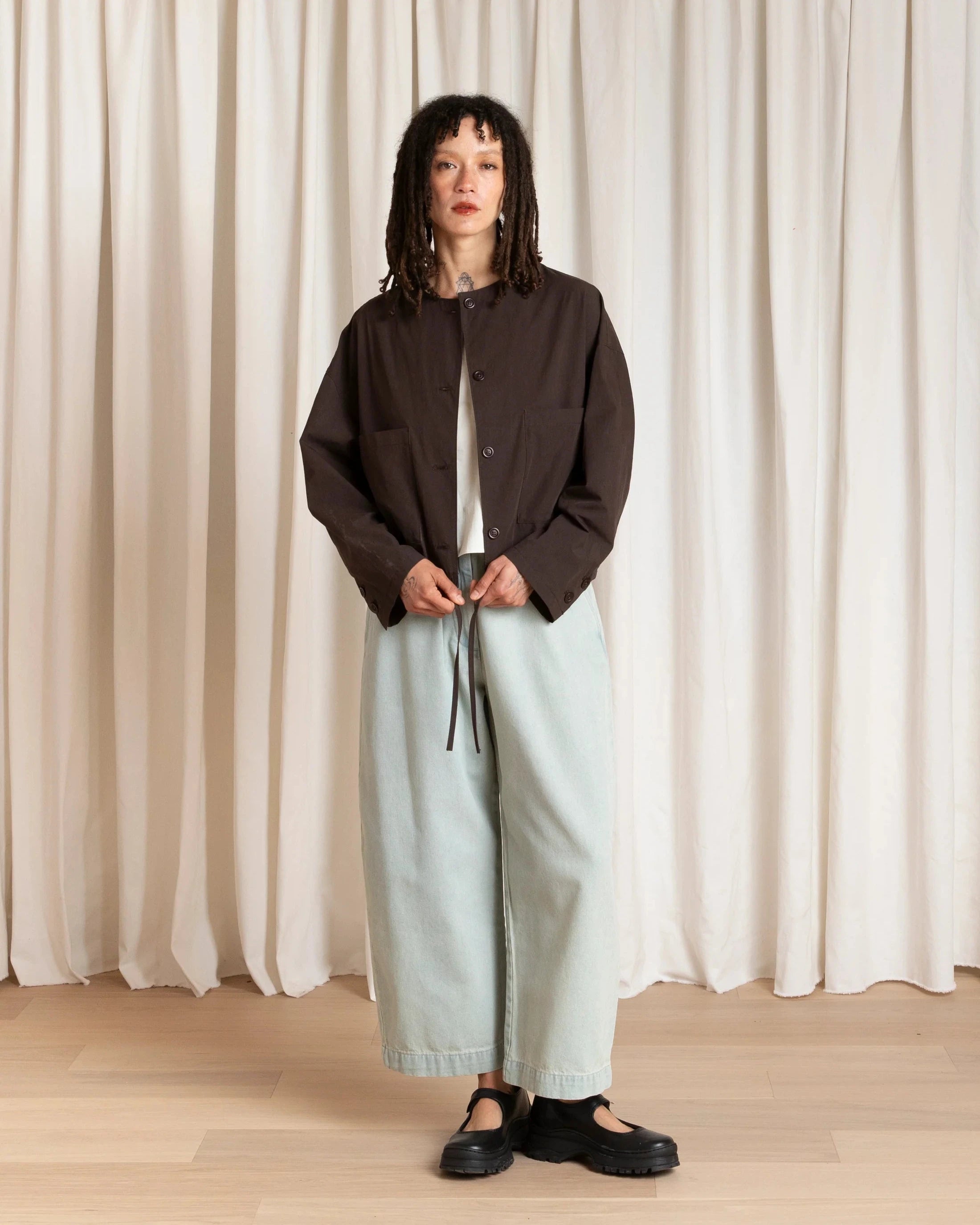 Person wearing a dark jacket and light pants standing against a white curtain.