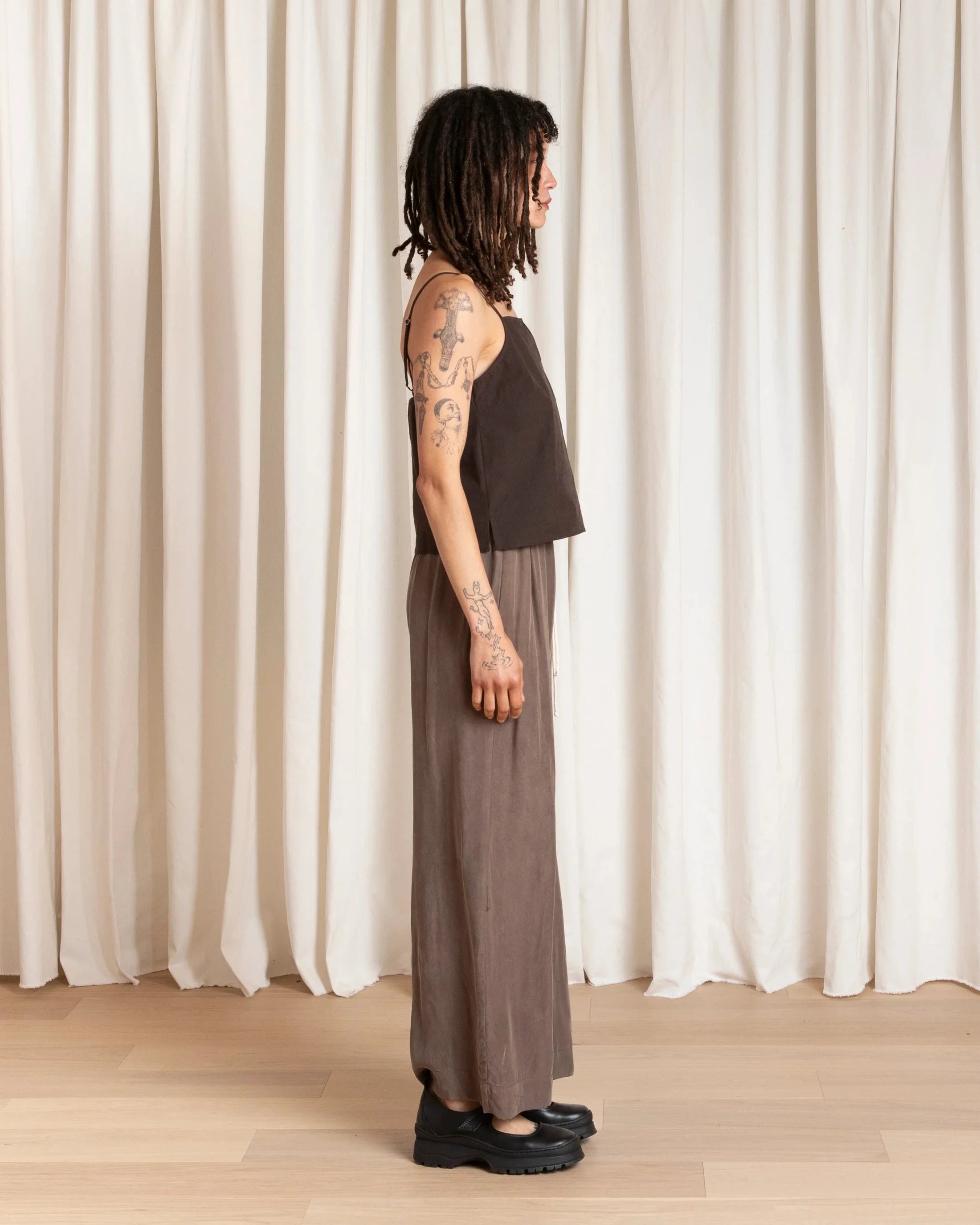 Person wearing a dark top and brown pants standing against a white curtain.