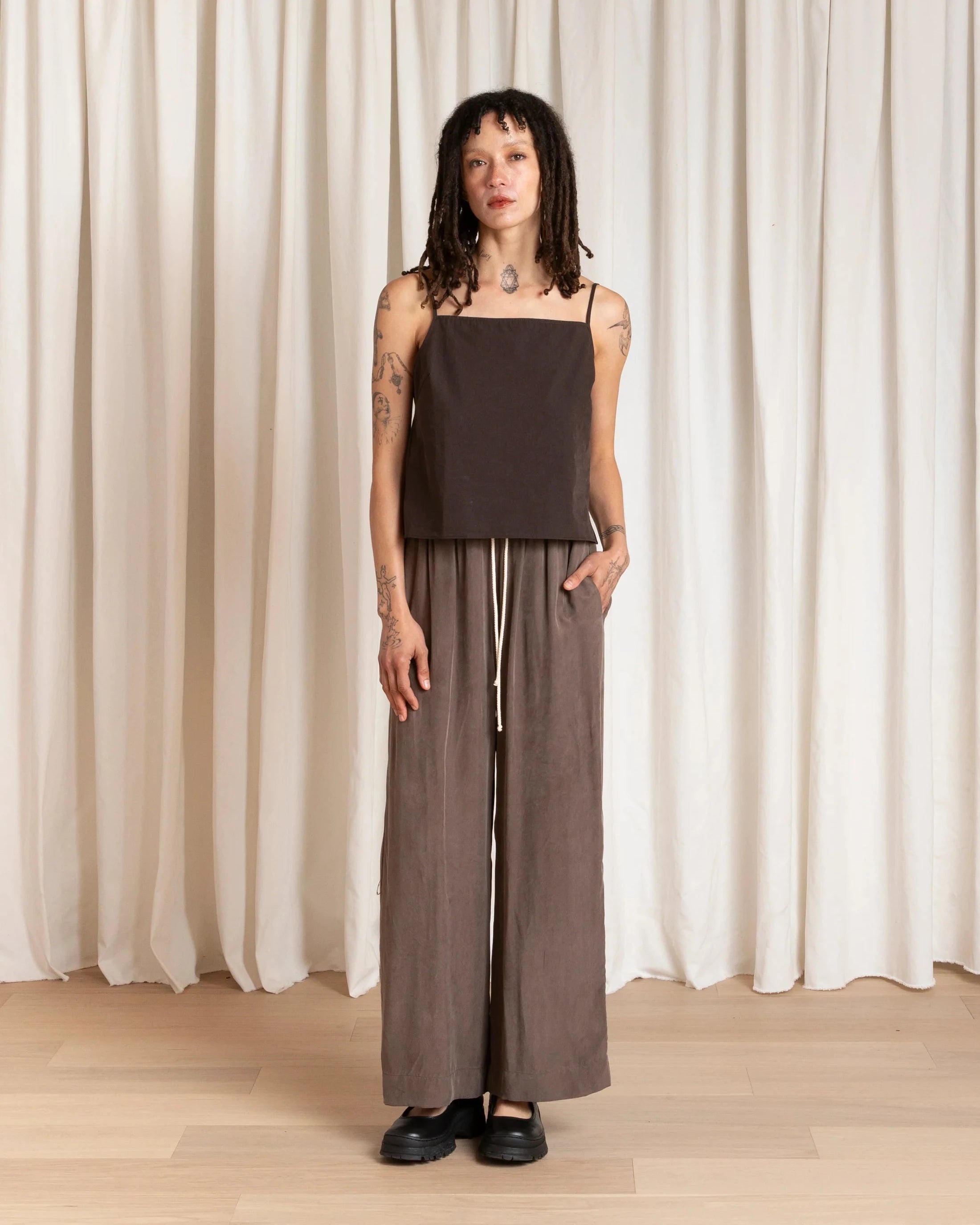 Person wearing a brown sleeveless top and wide-leg pants standing against a white curtain.