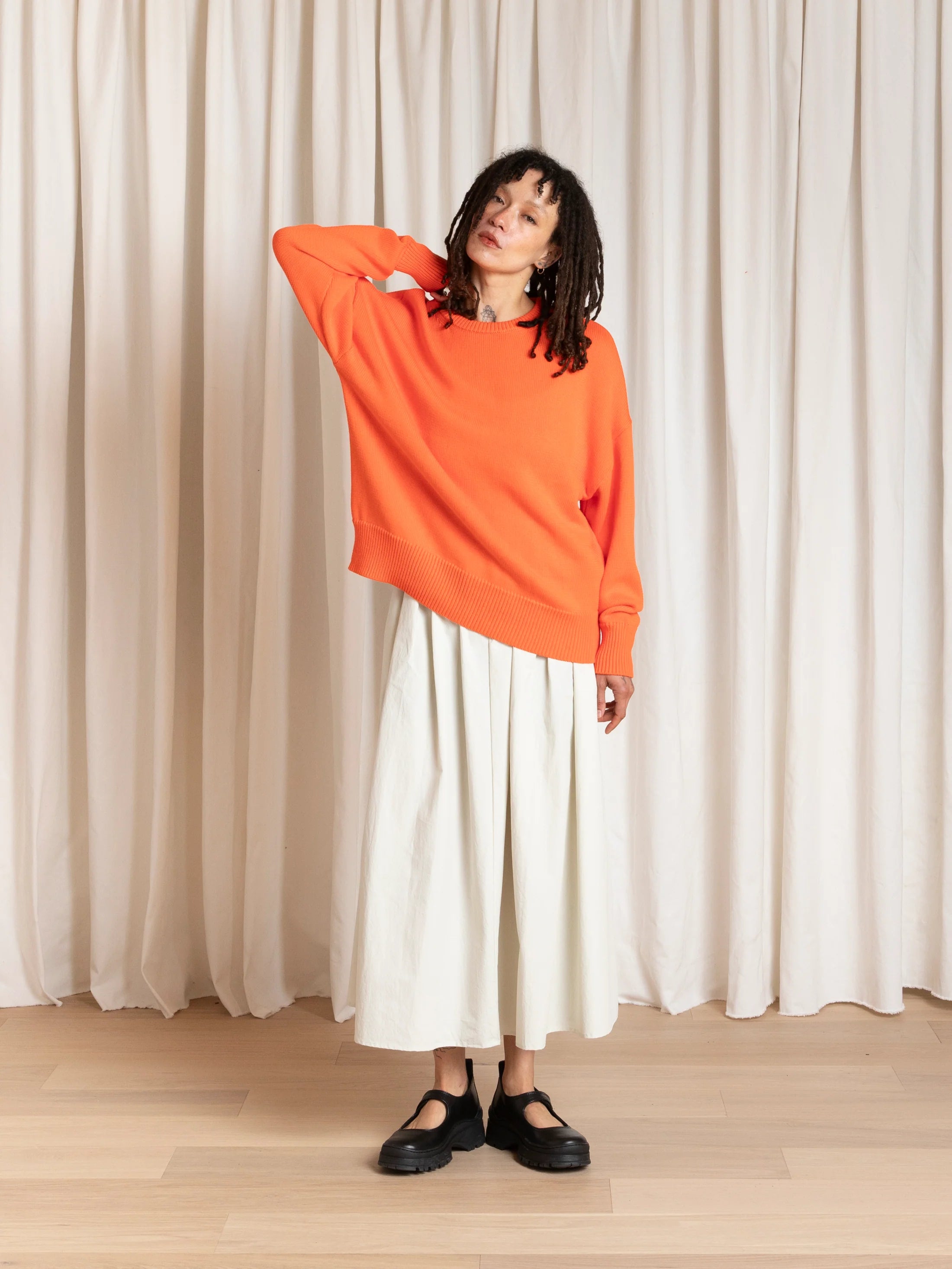 Person wearing an orange sweater and white skirt standing against a white curtain.