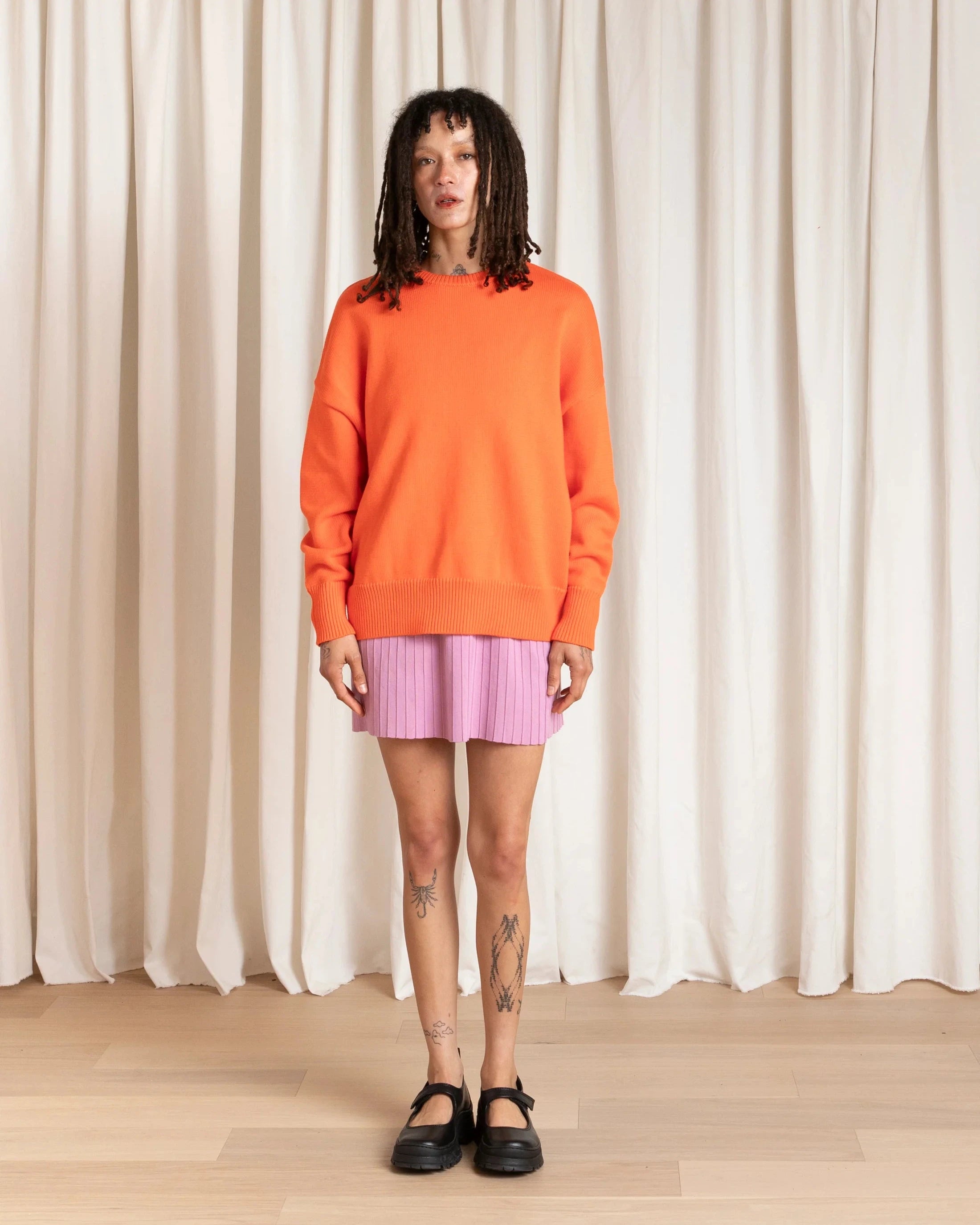 Person wearing an orange sweater and pink skirt standing against a white curtain.