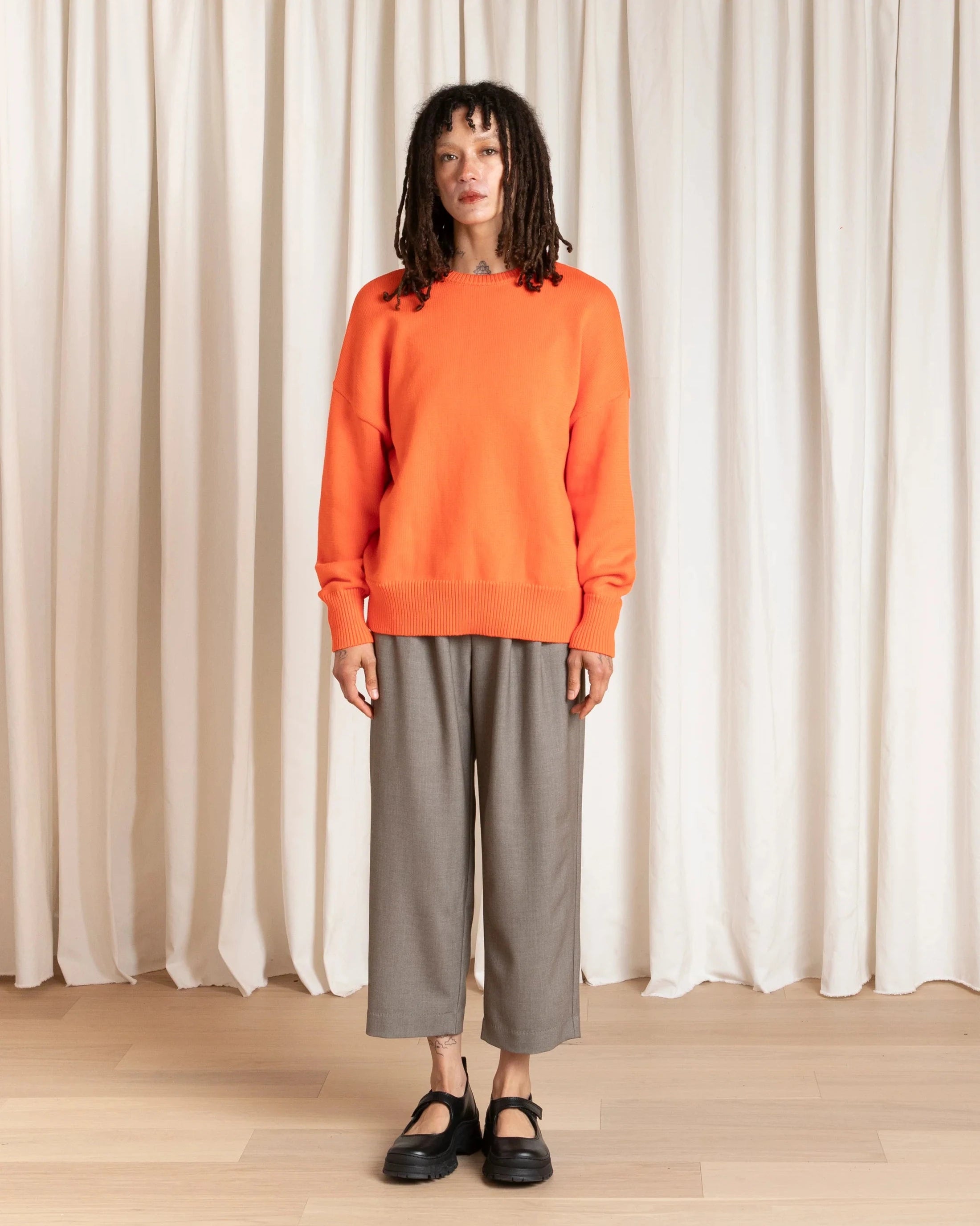 Person wearing an orange sweater and gray pants standing against a white curtain.