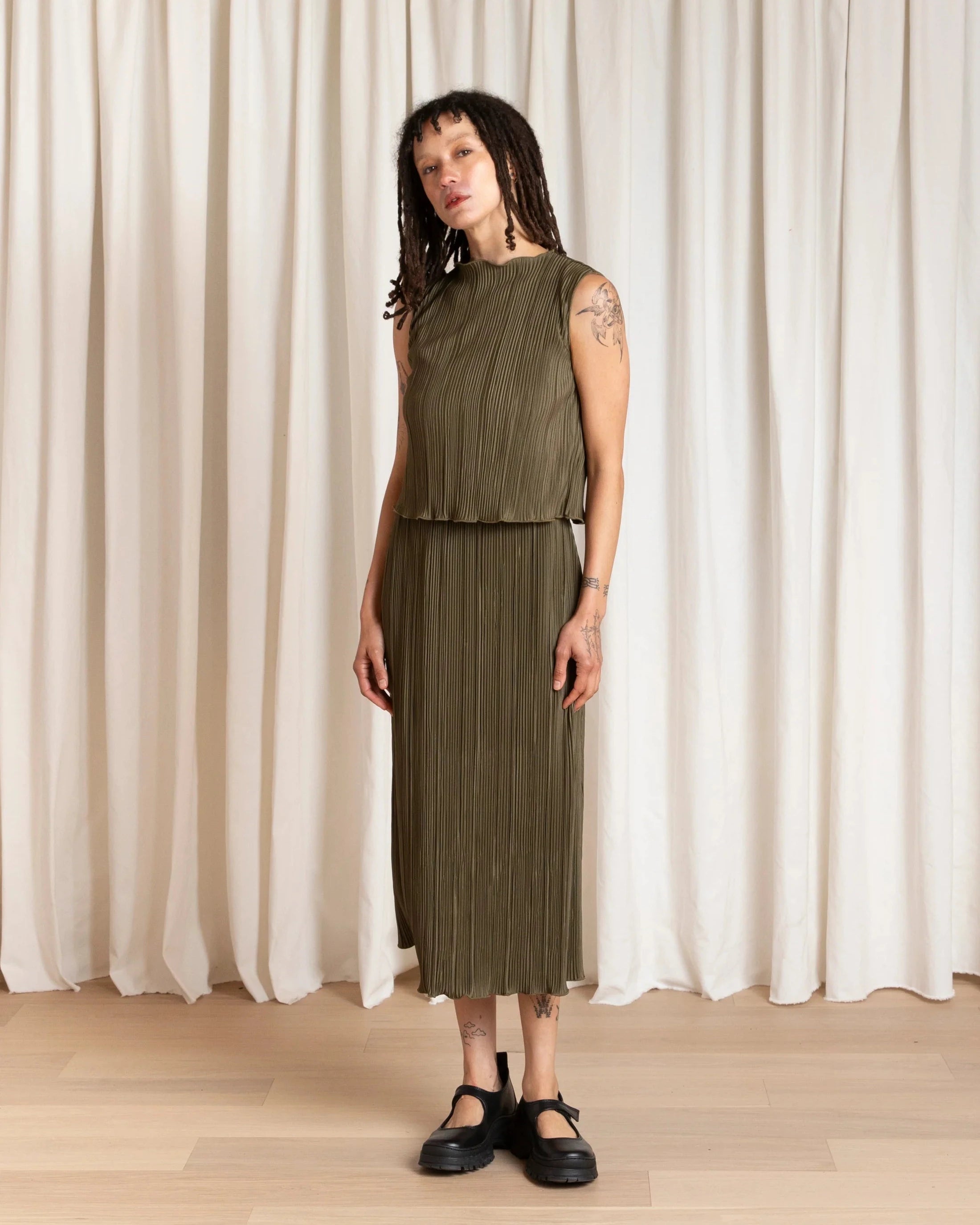 Woman wearing a green sleeveless top and pleated skirt standing against a white curtain.