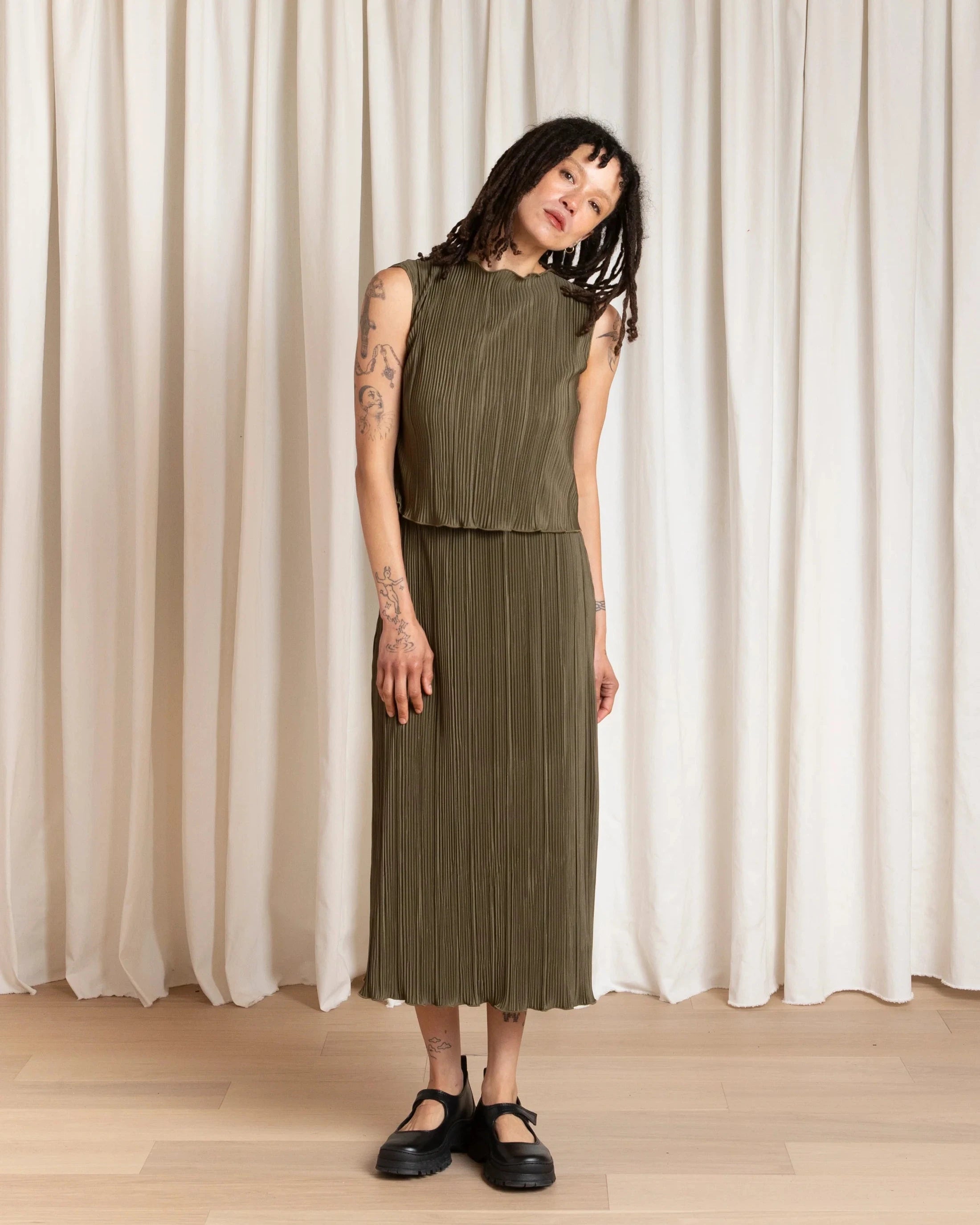 Woman wearing a green pleated dress standing in front of white curtains.
